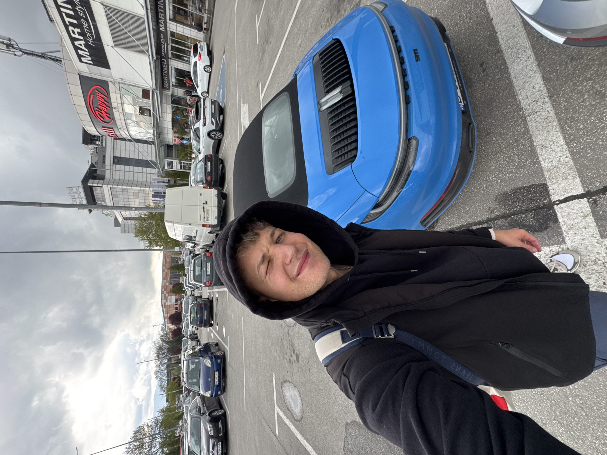 A selfie captured in a parking lot on an overcast day shows a young male subject winking and smiling at the camera. He wears a black hooded jacket with the hood up, and a blue strap, partially visible, displays the reversed text "YMMOT" (TOMMY), likely from a backpack. His sneakers, white and grey, are visible near the bottom right.
In the immediate foreground, to the right of the person, a bright blue Porsche 911 convertible is parked. Its rear engine cover and the "911" badge are clearly visible. Beyond this, the parking lot extends with numerous other vehicles, including a white Ford Kuga, a white van (license plate "PB 1720 HT"), a dark grey car (license plate "PB 2452 HA"), and a blue Toyota Yaris. The "PB" prefix on the license plates indicates Plovdiv, Bulgaria, aligning with the provided location context.
In the background, large commercial buildings are visible under the cloudy sky. One prominent building features multiple signs: "MARTI" and "MARTINELI Home style MARTIN" are seen on a white section, while a red, neon-style sign spells out "Happy." Another partial sign reads "apiter." The overall setting appears to be a large retail park or shopping center parking area. The ground is asphalt with white parking lines, suggesting a typical commercial parking environment.
