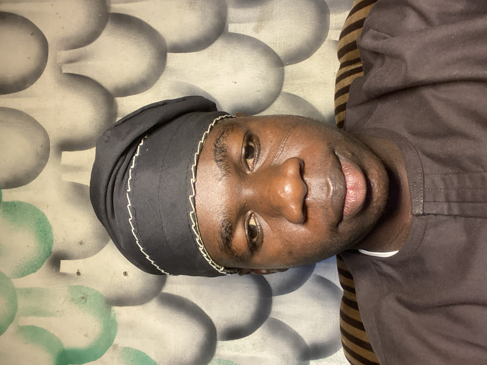 The image is a close-up, rotated portrait of an individual with dark skin, likely taken indoors. The subject, an adult, is oriented horizontally across the frame, with their head to the left and body extending to the right. They are looking directly at the camera with a neutral expression, eyes open and a slight parting of the lips.
The person is wearing a dark, possibly black, head covering or cap, which features a subtle white or light-colored embroidered edge or chain-like detail. They are also wearing a dark brown garment, possibly a shirt, visible on their upper body. Behind the individual's right shoulder, a striped pattern in shades of brown and lighter brown/beige is visible, suggesting a pillow or bedding.
The background consists of a textured or patterned surface. Large, rounded, bulbous shapes in shades of grey and white dominate the background, with some sections on the lower left exhibiting green or teal color splashes. This pattern could be a wall, wallpaper, or part of a mattress. The lighting appears even, suggesting an indoor environment without direct harsh sunlight. There is no visible text. Given the location context, the scene is likely in Mararaba, Nigeria, possibly within a home or personal dwelling.