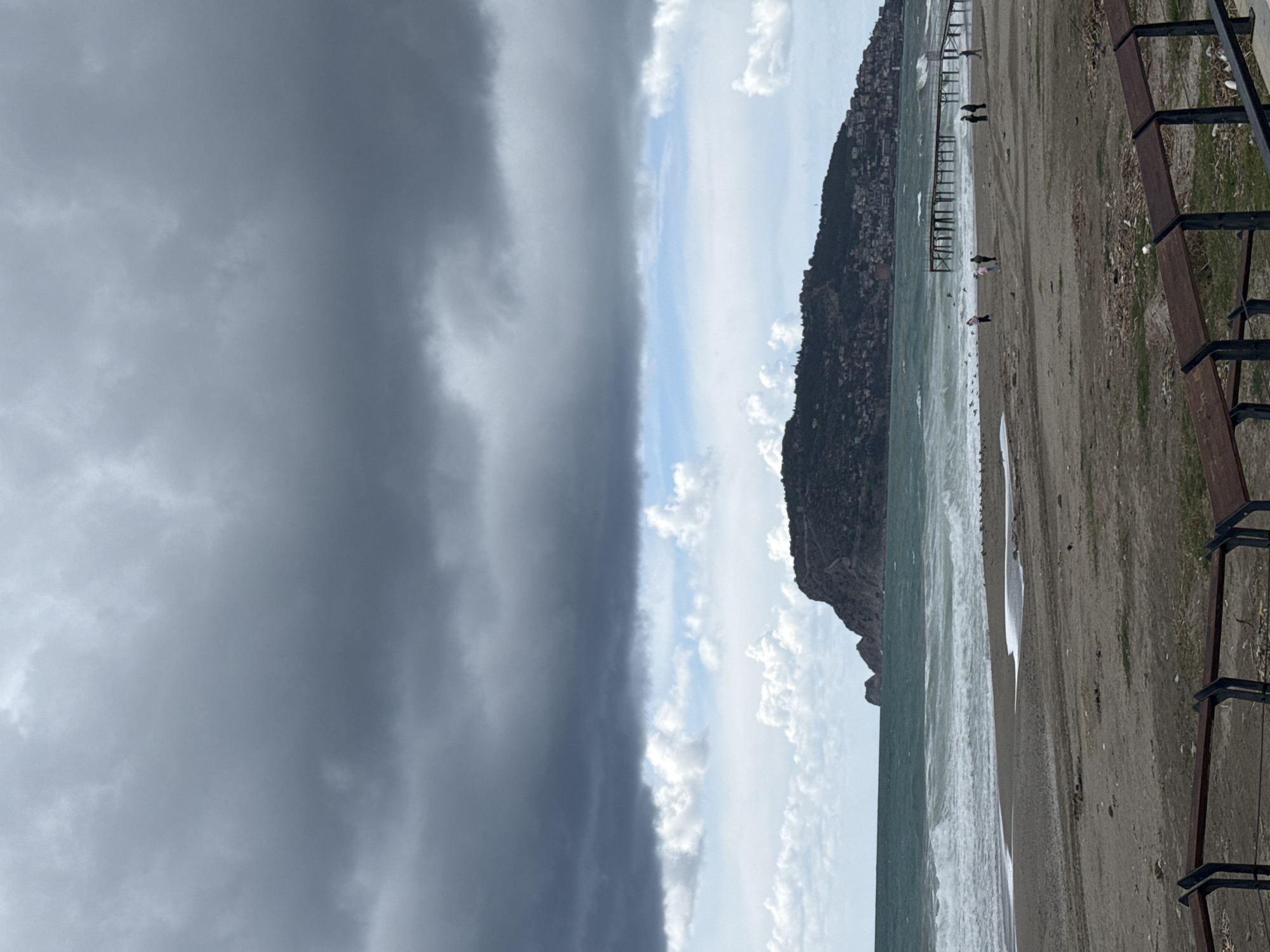 The image captures a beach scene in Alanya, Turkey, showing a stark contrast between an approaching storm and the coastline. The left side is dominated by a dense, dark cloud formation indicative of a storm, while the right side displays a lighter sky with scattered clouds. A mountain range with some residential development along its slopes is visible along the coastline. A pier extends out into the sea. The beach is sandy and there are some people on the shore. A fence runs along the edge of the beach. The sea is choppy with whitecaps.