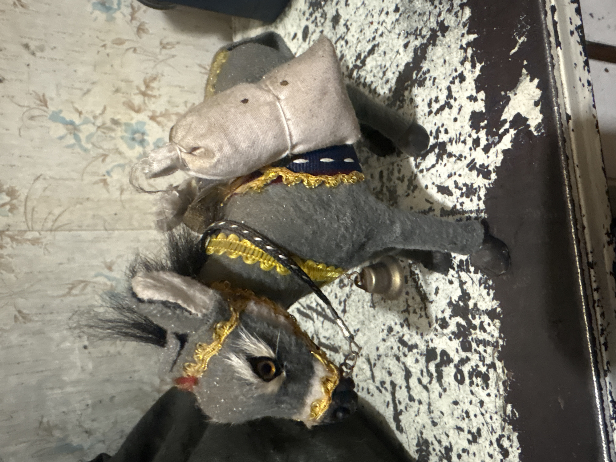 The image displays a vintage-style stuffed donkey toy positioned on an indoor surface. The donkey is predominantly gray, featuring a lighter muzzle and distinct amber-brown eyes. It has a shaggy black mane and tail. The toy is intricately adorned with gold braided trim around its head and body, and a dark blue and red sash is wrapped around its midsection. A light beige, sack-like object rests on its back, secured with a string, and a small metallic bell dangles from a chain near its head.
The background surface is divided. To the left, a faded light-colored wallpaper shows a delicate floral pattern with light blue and brown accents. To the right, the surface is heavily textured and distressed, featuring dark brown or black paint that has chipped away to reveal a lighter, possibly white or off-white, underlayer. This distressed area suggests an aged piece of furniture or an old wall, with a darker, more solid edge visible on the far right, implying a frame or border.
No specific activity or event is taking place; the image is a static portrayal of the toy. The lighting appears to be ambient indoor light. There is no visible text in the media file. The scene suggests an interior setting, possibly a home or an antique display, located in Krasnoyarsk, Russia.