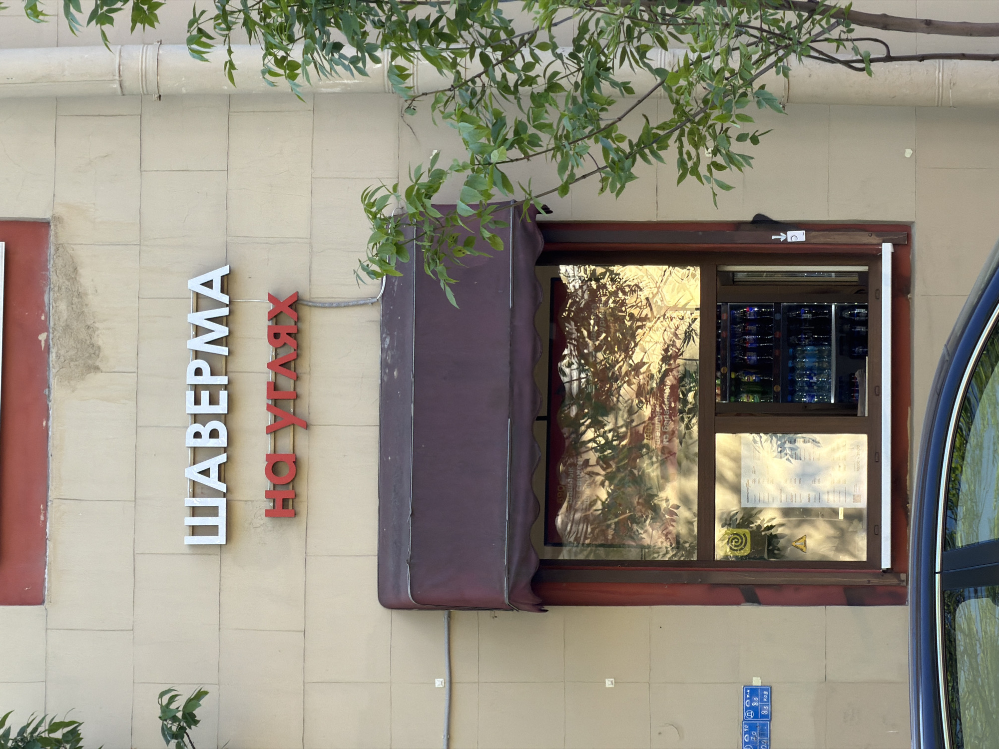 This image captures a section of a building facade in Saint Petersburg, Russia, likely belonging to a food establishment. The wall is made of light beige, rectangular tiles, with some signs of age and discoloration.
The primary subjects are a prominent vertical sign and a large window with an awning. The sign features two words in Cyrillic script. The upper word, in white block letters, reads "ШАВЕРМА" (SHAVERMA), indicating a shaurma or kebab shop. Below it, in red block letters, is "НА УГЛЯХ" (NA UGLYAKH), which translates to "on coals" or "charcoal-grilled," specifying the cooking method.
To the right of the sign is a window with a dark brown frame, partially covered by a maroon or dark purple retractable awning. The window glass is highly reflective, showing distorted reflections of green tree foliage and bright light, suggesting a sunny daytime. Through the reflections, the interior reveals what appears to be refrigeration units stocked with bottles, likely beverages. A white paper, possibly a menu or information sheet, is taped to the inside of the window, along with several symbols including an arrow pointing right, a circle, a yellow spiral, and a yellow warning triangle.
Overhanging branches with green leaves drape into the frame from the top, partially obscuring the upper wall and window. A beige drainpipe is visible along the top-left edge of the building. On the far right, the curved, dark blue body and window frame of a vehicle, likely a car, are visible, further reflecting the green surroundings. Electrical conduits and wires run along the wall beneath the window. The overall impression is a bustling urban setting with a commercial activity.