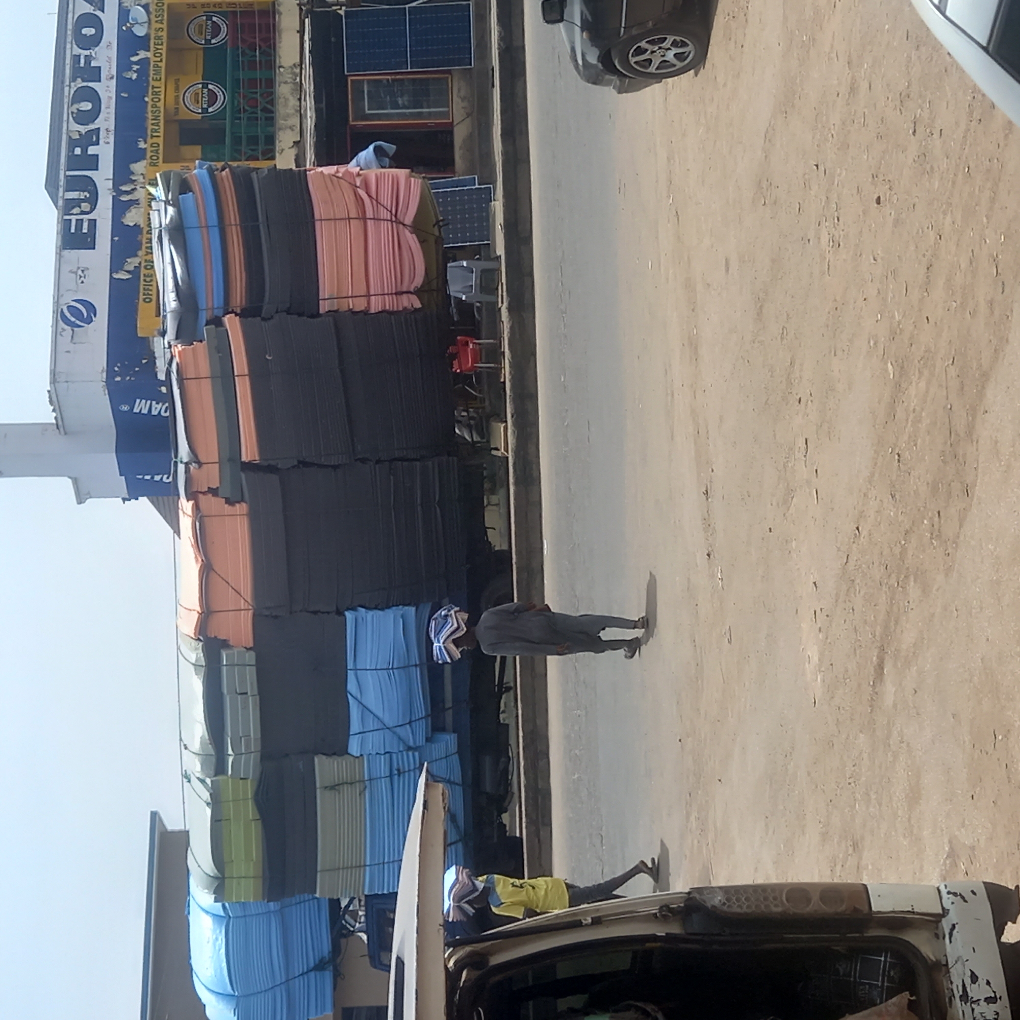 The image captures a bustling street scene in Dadi Riba, Nigeria, under bright daylight. The primary subjects include two individuals, large stacks of what appear to be foam mats or insulation panels, and several vehicles.
On the left side of the frame, a multi-story building features a large sign with "EUROFOA" prominently displayed in blue and white lettering, with smaller text "Flops og 34 Bould De" and "DAM". Below this, another sign in yellow and green reads "OFFICE OF YAMEL ROAD TRANSPORT EMPLOYER'S ASSO" and "RTEAN" repeated twice, with "YAM AVIA CRAPE" visible between. The building also has windows with what look like solar panels mounted on the exterior.
Along the street, massive stacks of foam mats in various colors—black, pink, blue, light green, and grey—are piled high, taking up a significant portion of the scene. These stacks are tied together, suggesting they are ready for transport or storage.
Two men are visible in the foreground. One man, dressed in dark clothing with a striped head covering, walks along the dusty ground next to the stacked materials. Another man, wearing a yellow shirt and dark trousers with a similar head covering, is near the back of a white truck or van, possibly engaged in loading or unloading items. Parts of a dark-colored car are visible at the top and bottom of the frame. The ground is a mix of dirt and paved road. The bright sun casts clear shadows, indicating midday conditions. The overall impression is a commercial area dealing with building or insulation materials.