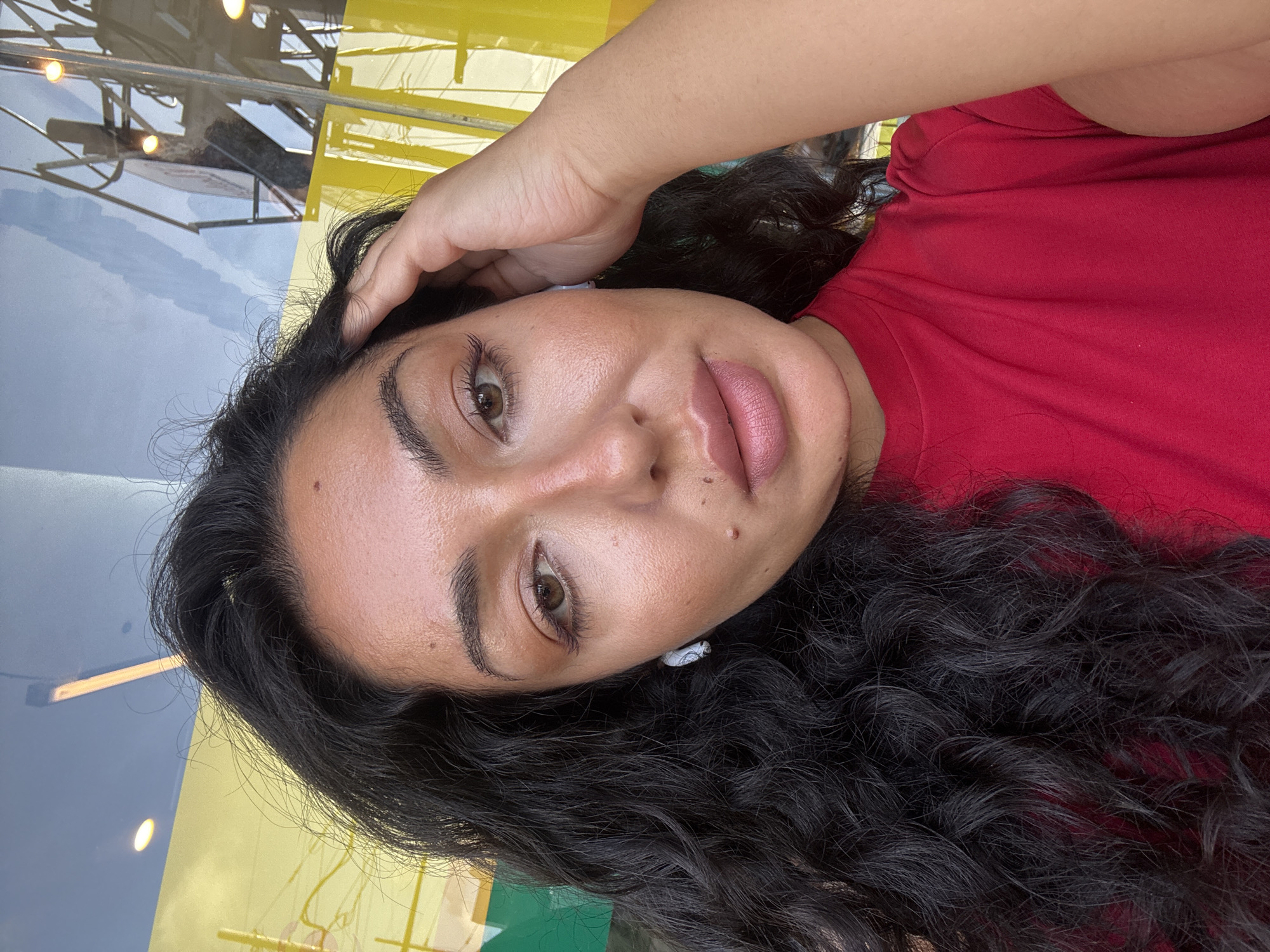 A close-up, rotated portrait of a woman with dark curly hair wearing a red shirt. She is holding her hand to her head. Her skin tone is medium, and she has brown eyes. She appears to be wearing light makeup, specifically lipstick. The background contains reflections of a structure with yellow and blue elements. Based on the environment, it's difficult to confirm the location within Kuta Selatan, Indonesia without additional visual cues.