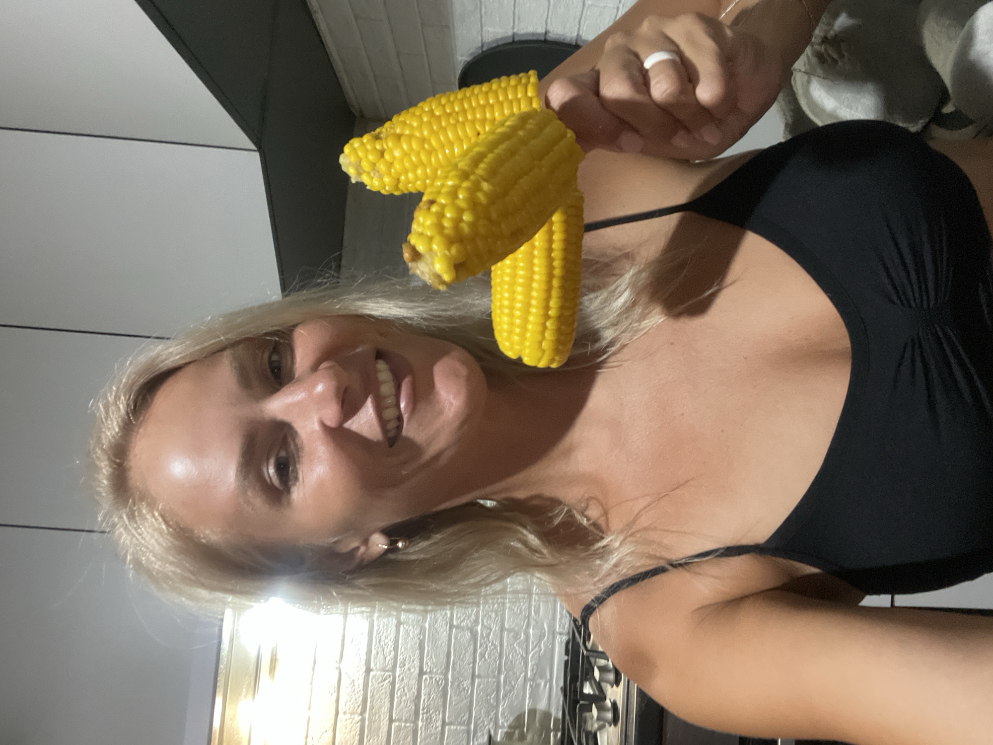In a kitchen setting, a woman with blonde hair and fair skin is holding up two ears of cooked corn, presenting them to the camera. She is wearing a black tank top and a white ring on her right ring finger. The background shows a white brick wall and kitchen appliances. There is no indication of the specific city of Baranovka, Russia.