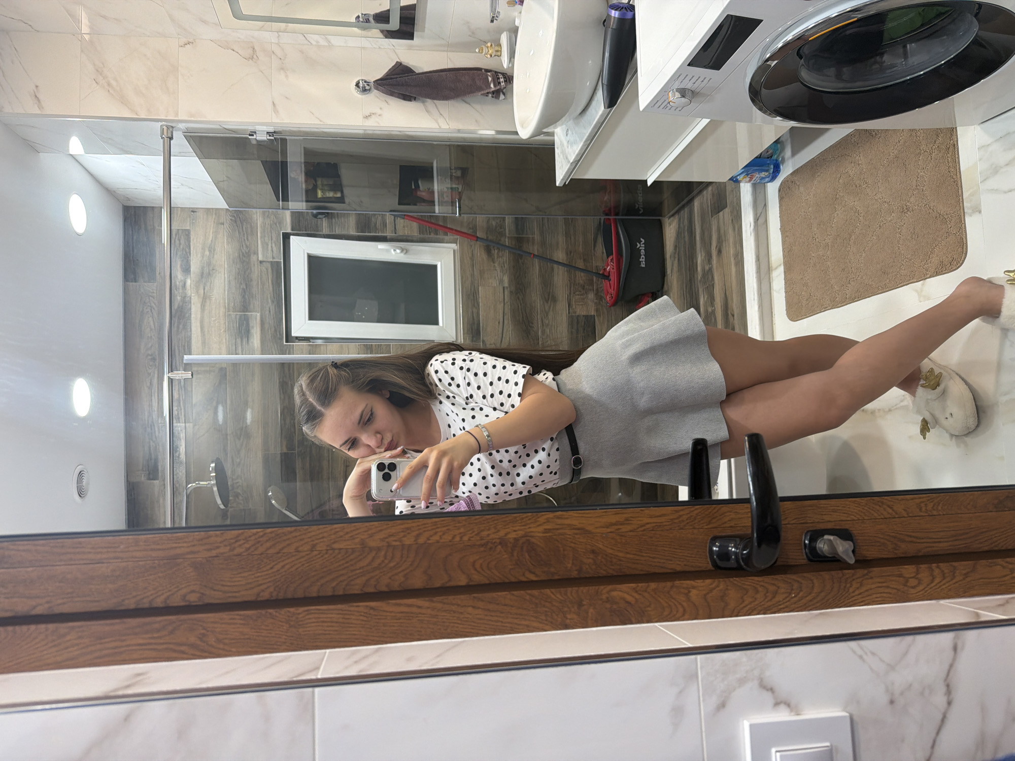 A girl takes a selfie in a bathroom with a washing machine, sink, and mop. The bathroom has light brown and white tiles. The girl is wearing a white polka dot shirt and a grey skirt. It is not possible to confirm the location as Buhovo, Bulgaria, based on the image.