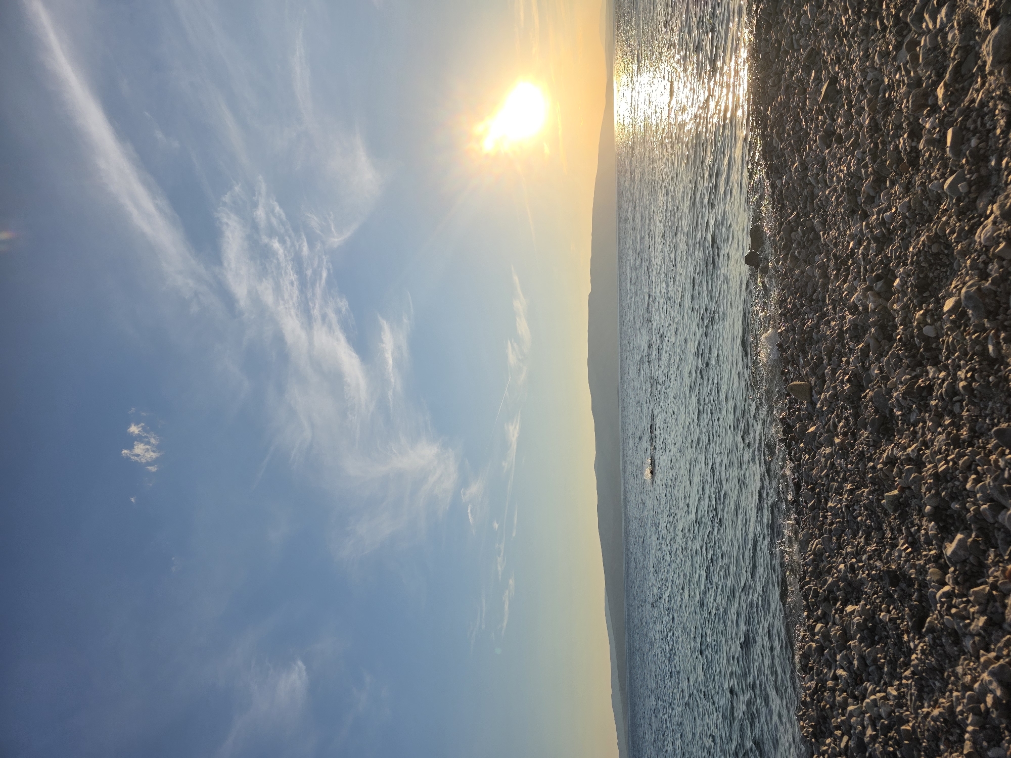 A low-angle, wide shot captures a serene beach scene at dusk. The foreground is dominated by a dense scattering of smooth, grey pebbles, some of which are wet from the receding tide. The ocean, a deep blue, stretches out to the horizon, its surface rippled by gentle waves that lap against the shore. In the distance, a long, low mountain range, silhouetted against the sky, forms a muted backdrop. The sun, a bright, radiant orb, hangs in the upper right quadrant of the image, casting a warm, golden glow across the water and illuminating the sky with streaks of white and pale yellow clouds. The overall atmosphere is one of tranquility and natural beauty.
