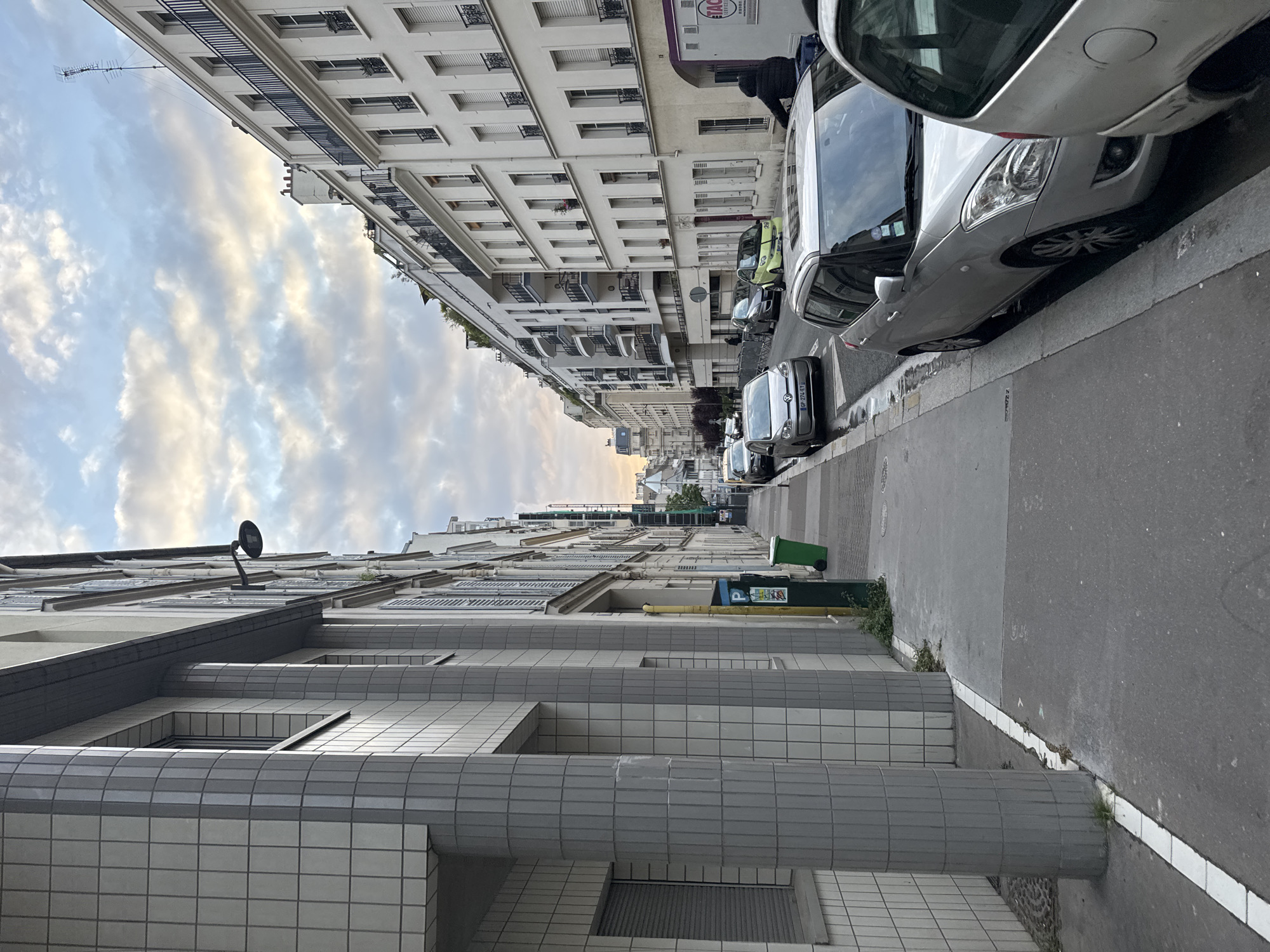 The image shows a street in Paris, France, lined with buildings on both sides. Cars are parked along the right side of the street. The buildings are predominantly white or light-colored, with multiple stories and windows. A street lamp is visible attached to the lower building. The sky is cloudy. A green dumpster is positioned on the side of the road. The street is asphalt. The image is taken at an angle, creating a perspective view down the street.