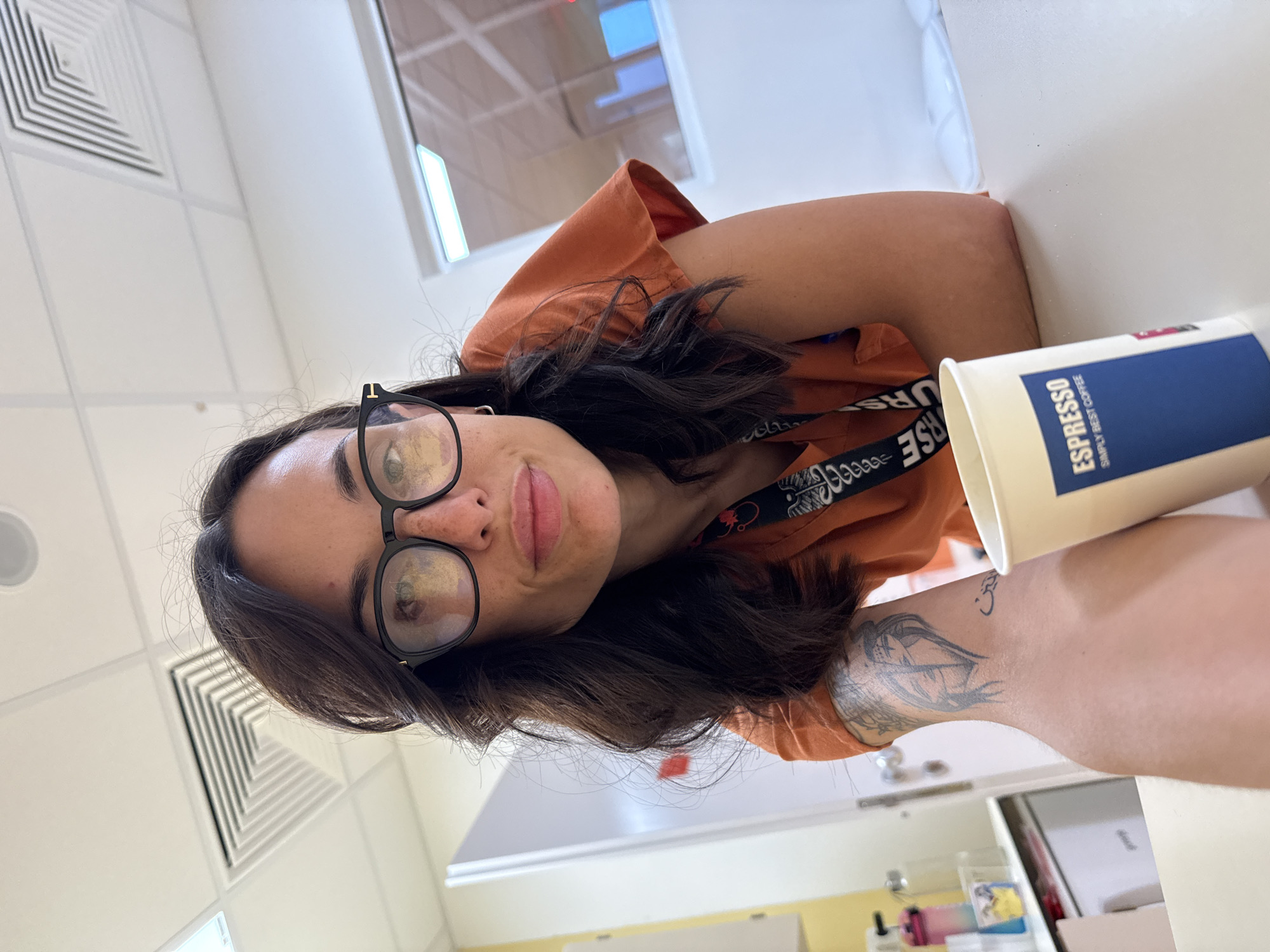 A woman with dark hair and glasses is shown in a selfie. She is wearing an orange shirt and a lanyard with a black and white design. She is holding a disposable coffee cup with "ESPRESSO" printed on it. Her left arm has a tattoo. The background appears to be an office setting with a white wall, vent, and window. The location in the city of Pleven, Bulgaria cannot be determined from the information presented.