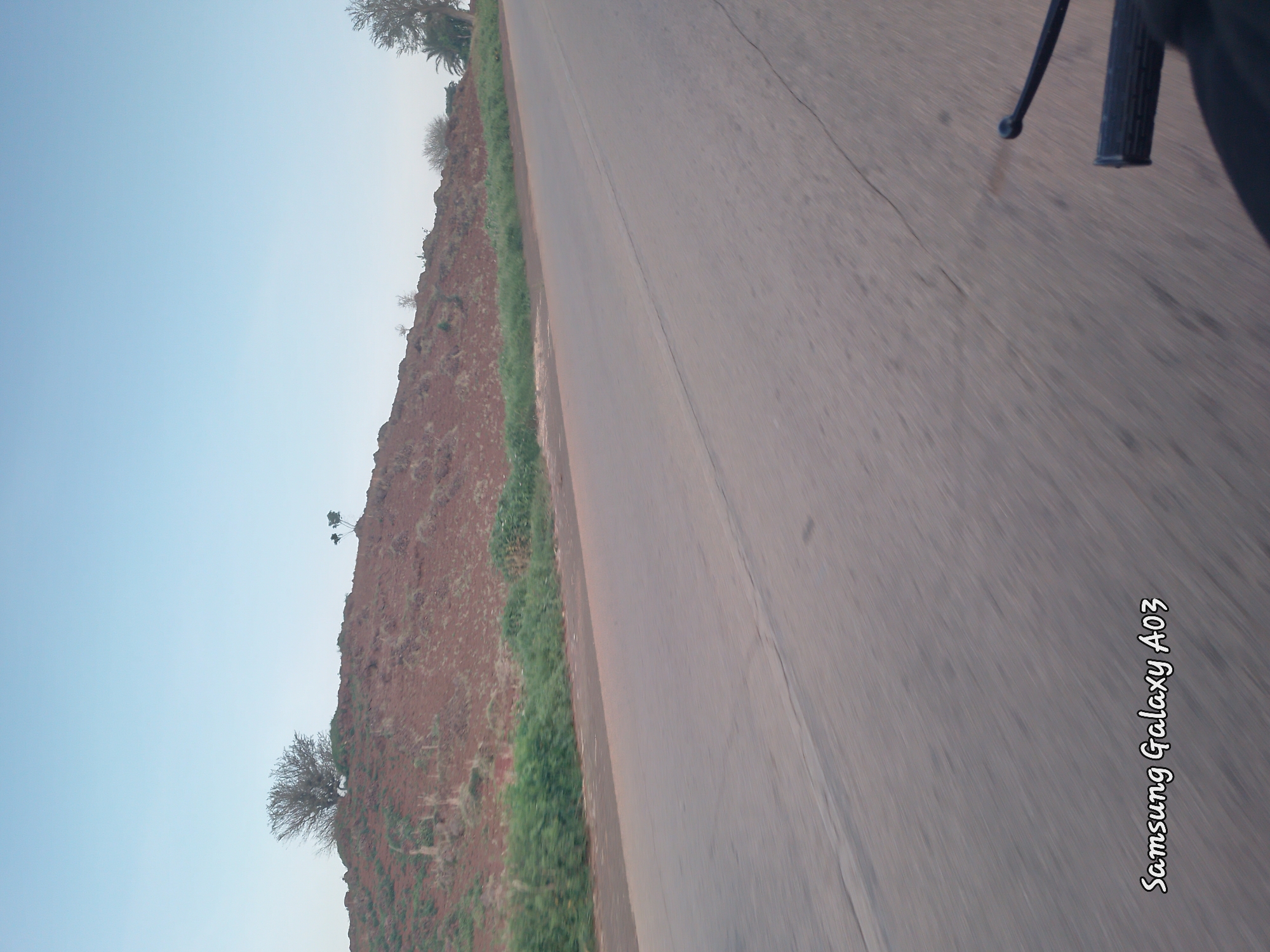 This image was captured from a moving vehicle, likely a motorcycle given the visible handlebars in the lower right corner, on a paved road. The road runs along the base of a gently sloping, reddish-brown hill covered in sparse vegetation and a few scattered trees. The sky above is a clear, pale blue. In the lower left corner of the image, white text reads "Samsung Galaxy A03", indicating the device used for capture. The overall scene suggests a rural or semi-rural environment in Doka-Tofa, Nigeria, possibly during daylight hours with fair weather. There are no people visible in the frame, and the focus is on the landscape and the road.