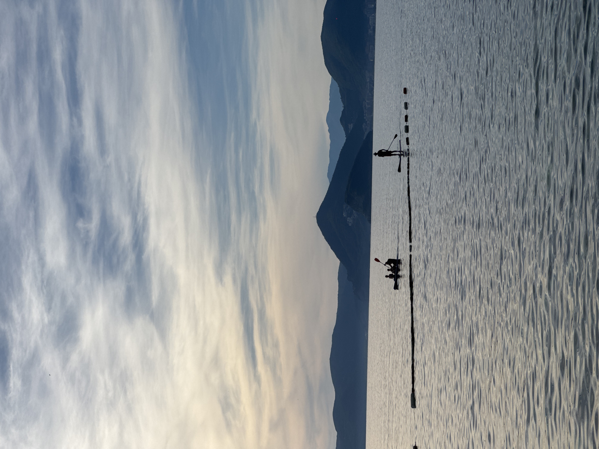 The image depicts a calm body of water with silhouetted figures engaged in water activities against a backdrop of mountains. In the mid-ground, a small boat, possibly a kayak, carries at least three individuals. To the right, a single person stands on a paddleboard, holding a paddle. A line of buoys is visible in the water near the paddleboarder. The water is gently rippled, reflecting the soft light of the sky. The horizon is marked by a range of dark, undulating mountains, some with faint signs of human settlement. The sky is filled with wispy clouds, transitioning from a pale yellow near the horizon to a soft blue higher up. The overall impression is one of tranquility and outdoor recreation in a coastal setting. The image appears to be taken in the vicinity of Rijeka Reževići, Montenegro.