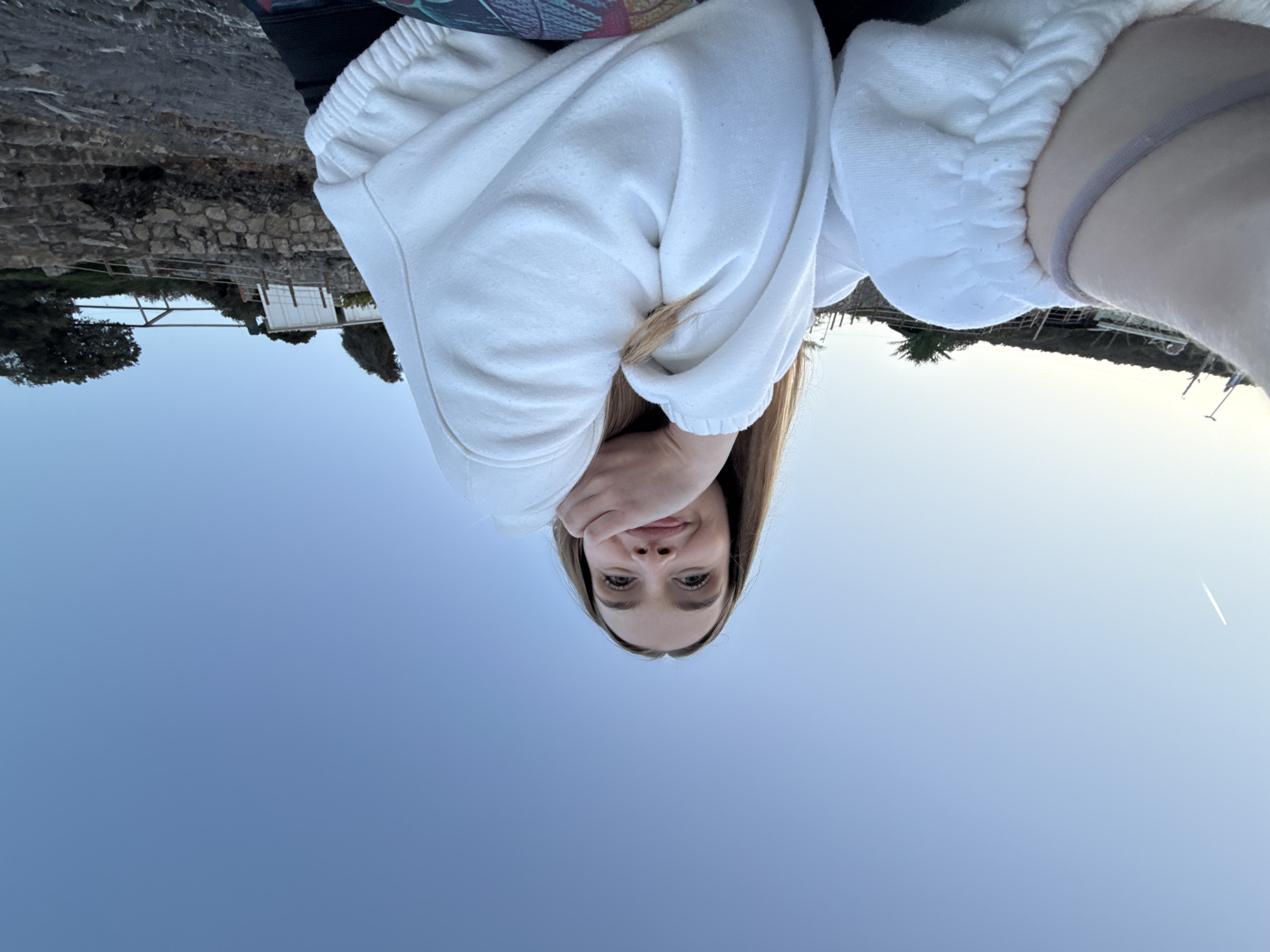 This full-shot selfie captures a young woman looking directly at the camera, holding the device from a slightly lower angle. She has long, straight light brown hair, and her left hand rests beneath her chin, creating a relaxed pose. She wears a white, possibly terry cloth, hooded sweatshirt or jumper, with a dark garment visible near her left shoulder. A grey elastic band is on her right wrist. Her expression conveys a subtle smile and a sense of contentment.
The setting appears to be a coastal or lakeside area in Seoca, Montenegro. Behind the woman, a textured stone wall or rocky terrain is visible, interspersed with dark green foliage and bushes. In the midground, beyond the wall, a distant shoreline stretches across the frame, featuring small white buildings, possibly houses or sheds, and more trees. On the far right, faint structures resembling docks or boats can be seen along the water's edge.
The clear sky dominates the upper portion of the image, transitioning from a deeper blue near the horizon to a lighter, almost white-blue towards the top. A faint, thin white contrail, likely from an airplane, is visible in the upper right quadrant of the sky. The lighting suggests either early morning or late afternoon, with soft, even illumination and no harsh shadows, indicating calm and pleasant weather. There is no visible text in the image.