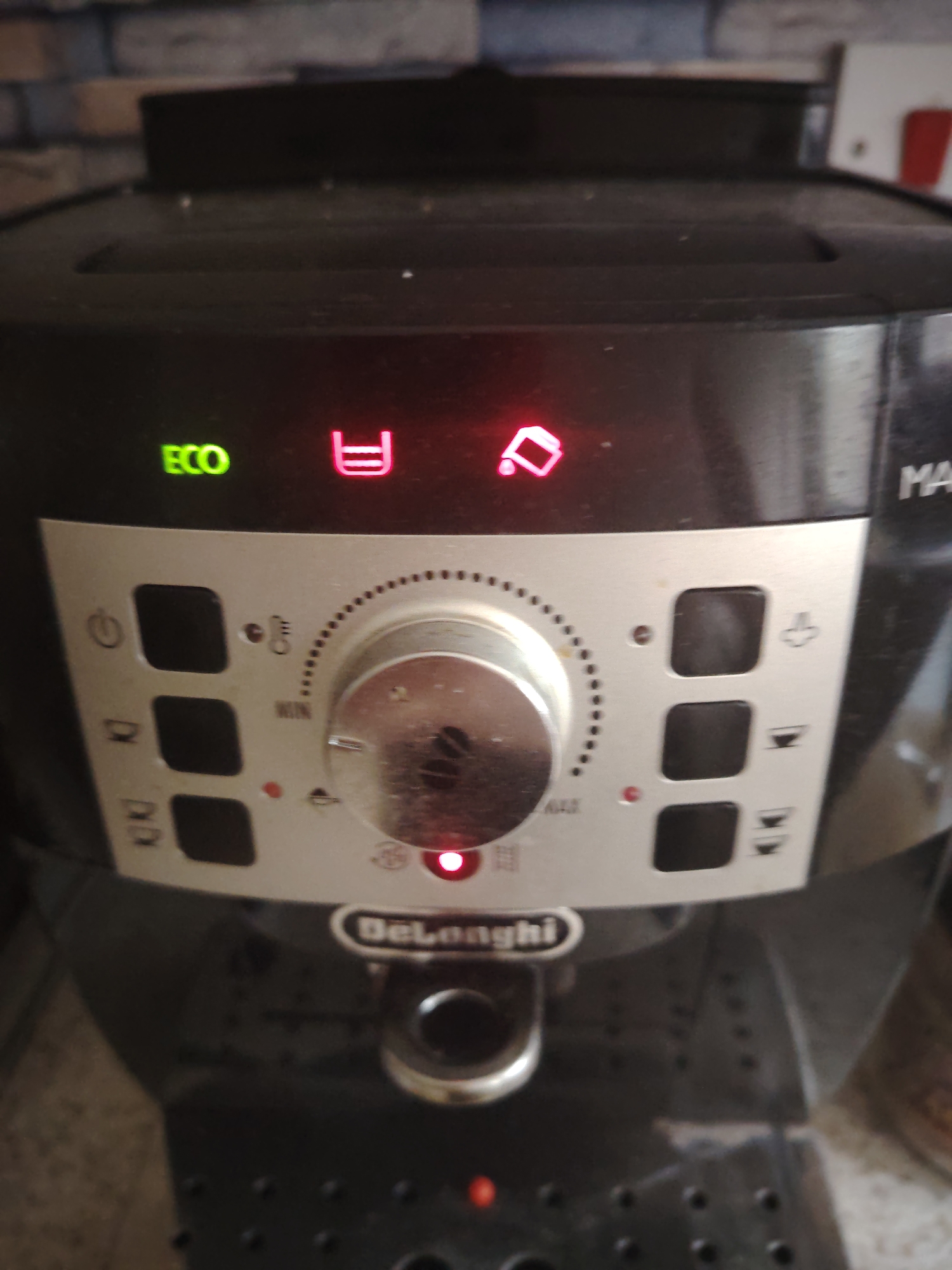 An image shows a close-up view of a black DeLonghi coffee machine. The machine features several indicator lights and controls. A green "ECO" light is illuminated on the left side. To its right, a red indicator light shaped like a full cup is on, and next to that, a red indicator light depicting a partially full cup with a drop falling from it is also illuminated. Below these lights is a silver dial with markings for "MIN" and "MAX" and dots indicating settings. To the left of the dial are three black buttons, and to the right are three more black buttons, each with an icon. A red indicator light is visible below the dial. The DeLonghi logo is printed in white on a black band above the drip tray. The background consists of a tiled wall. There is no indication of a specific location within Huntingdonshire, United Kingdom, in the provided image.