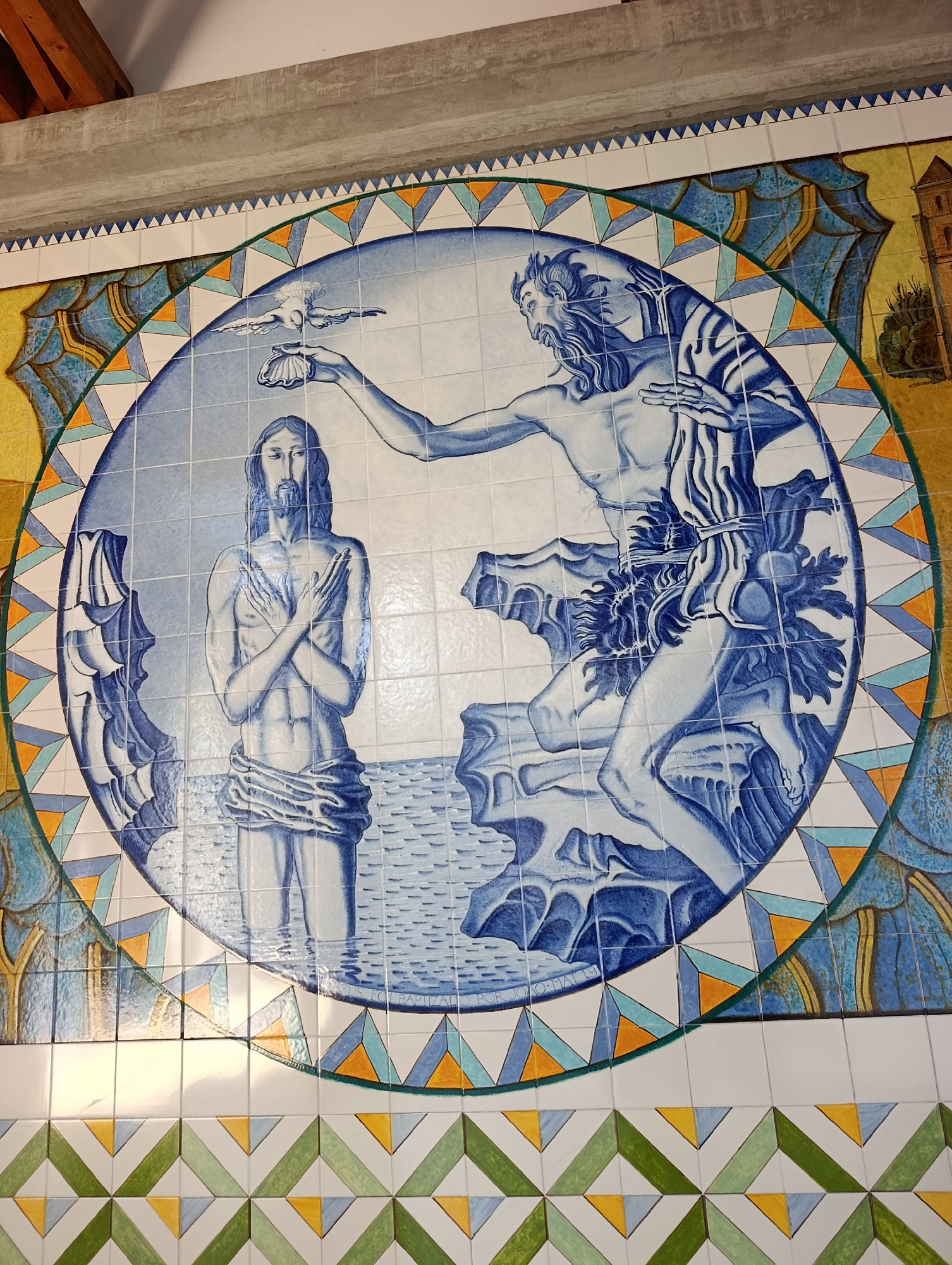 This image depicts a ceramic tile mural in Terras de Bouro, Portugal, likely a religious scene, given the imagery. The central focus is a circular design featuring two figures. On the left, a man with long hair and a beard stands with his arms crossed over his chest, appearing to be submerged in water. On the right, another man, also bearded and muscular, is depicted with his arm extended, holding a shell from which a dove is descending. This scene strongly suggests the baptism of Jesus by John the Baptist.
The figures are rendered in shades of blue and white, typical of azulejo tile art. The circular motif is bordered by a decorative band composed of triangular patterns in yellow, orange, and blue, with green accents. The background outside the circle features elements of architectural structures and natural landscapes, also rendered in ceramic tiles.
Below the main circular mural, there is a horizontal band of patterned tiles featuring a repeating geometric design of green and white chevrons with yellow triangular accents. The overall impression is of a vibrant and detailed public art installation. The specific location is likely a public building or a prominent wall in the town, given the scale and decorative nature of the artwork.