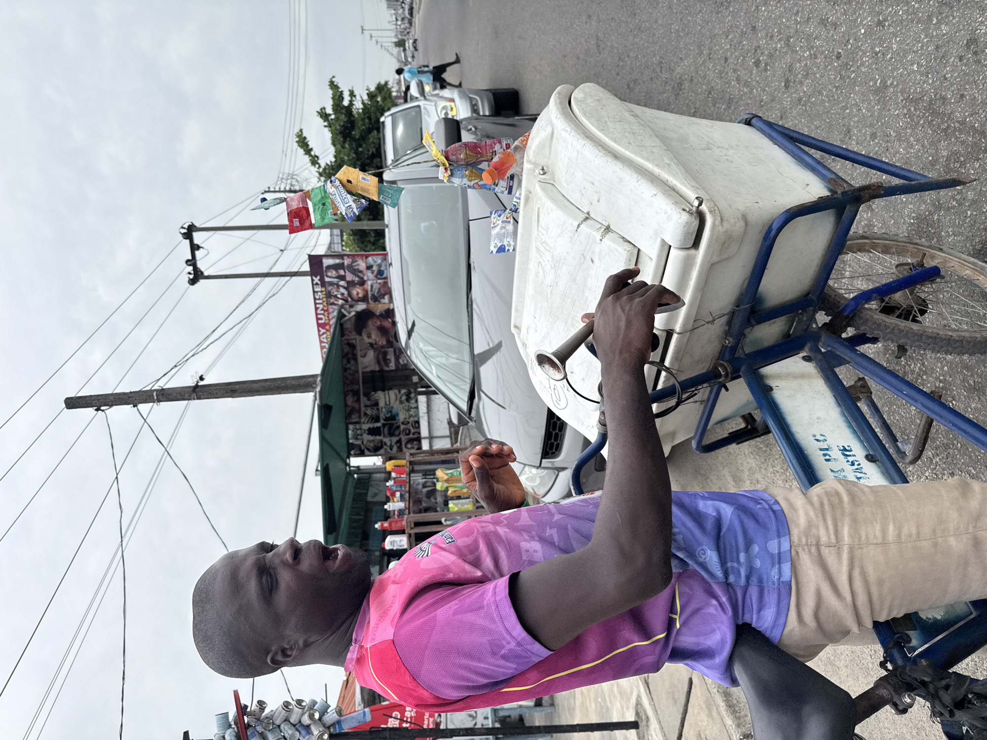 A man is riding a bicycle with a cooler mounted on the front in Ado, Nigeria. He is wearing a purple and pink t-shirt and beige pants. He is gripping a metal handlebar attached to the cooler. The cooler is white. The bicycle is blue with a white plate displaying blue text. Ado, Nigeria is visible in the background with cars, shops, power lines, and people along the street. A beauty salon is visible with a sign displaying various hairstyles. Bags of product hang from the bike.