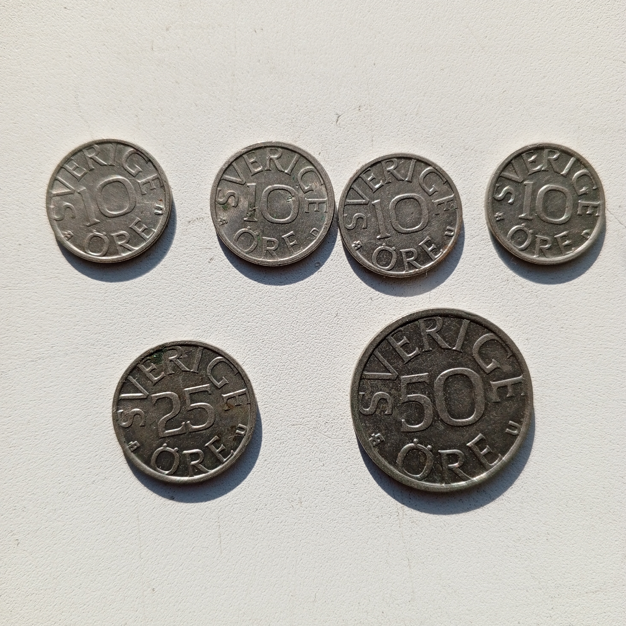 The image displays six Swedish coins laid out on a textured white surface, presumably a table or countertop. The coins are arranged in two rows. The top row contains four coins, each appearing to be a 10 öre denomination, with "SVERIGE" inscribed around the edge and "10 ÖRE" in the center. The bottom row features two coins: one is a 25 öre coin, and the other is a 50 öre coin. These coins are also inscribed with "SVERIGE" and their respective denominations. The lighting casts distinct shadows from the coins onto the surface, indicating a light source from the upper left. There are no people or city landmarks visible in the image. The scene is a close-up view of the coins. The location in Sayanogorsk, Russia, is not discernible from the provided image.