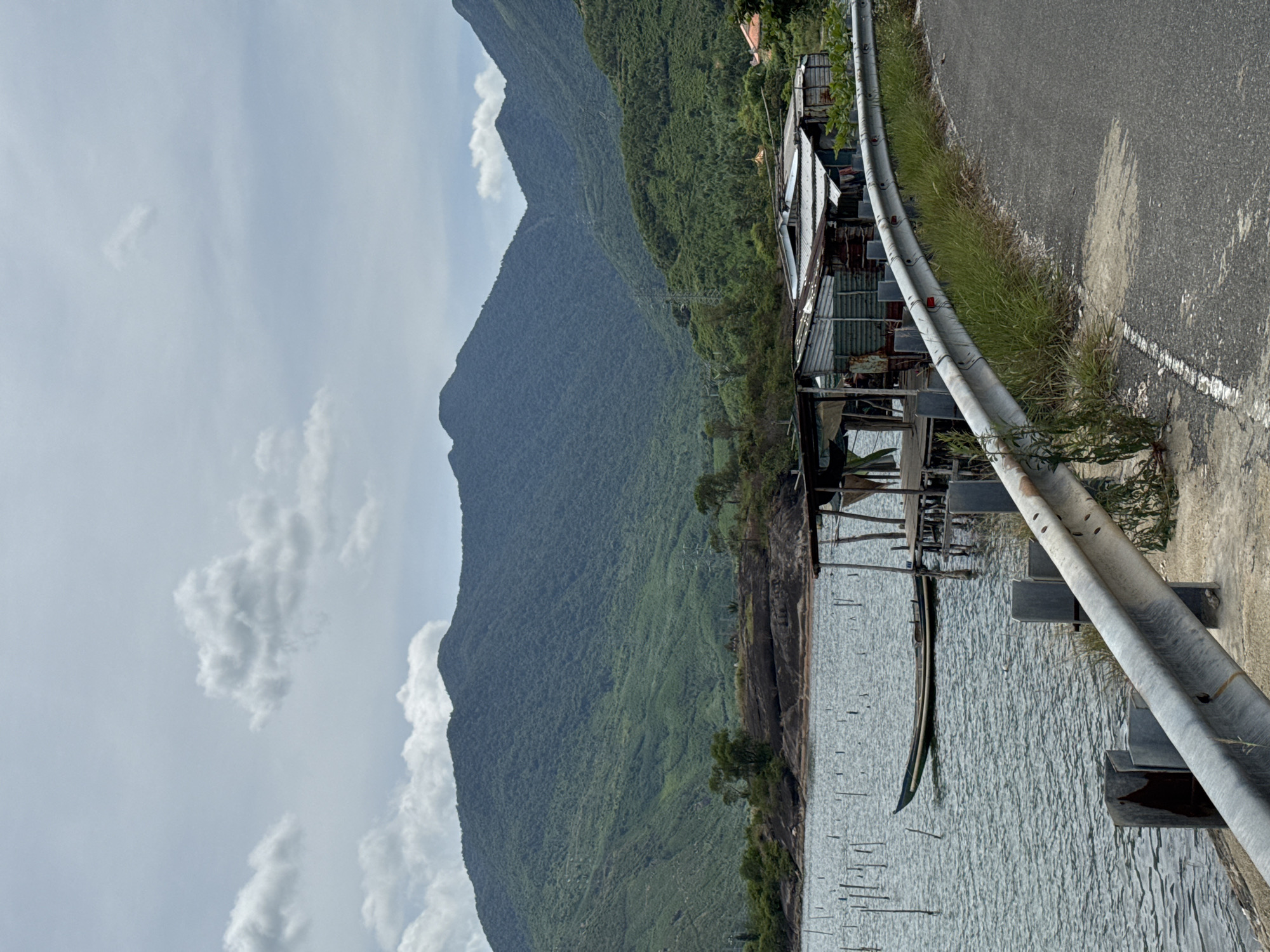 The image depicts a scenic view of a body of water with mountains in the background, likely on the outskirts of Huế, Vietnam. A single, narrow boat is moored on the water, near a rustic wooden stilted structure that appears to be a dwelling or a fishing platform. The structure is made of corrugated metal and wood, with tarpaulins providing additional cover. A paved road with a metal guardrail curves into the foreground, bordered by grass. The water is calm with ripples, and numerous vertical posts stick out of it, possibly marking a fishing area or aquaculture. The mountains in the distance are covered in lush green vegetation. The sky is overcast with scattered clouds. There are no people visible in the frame.