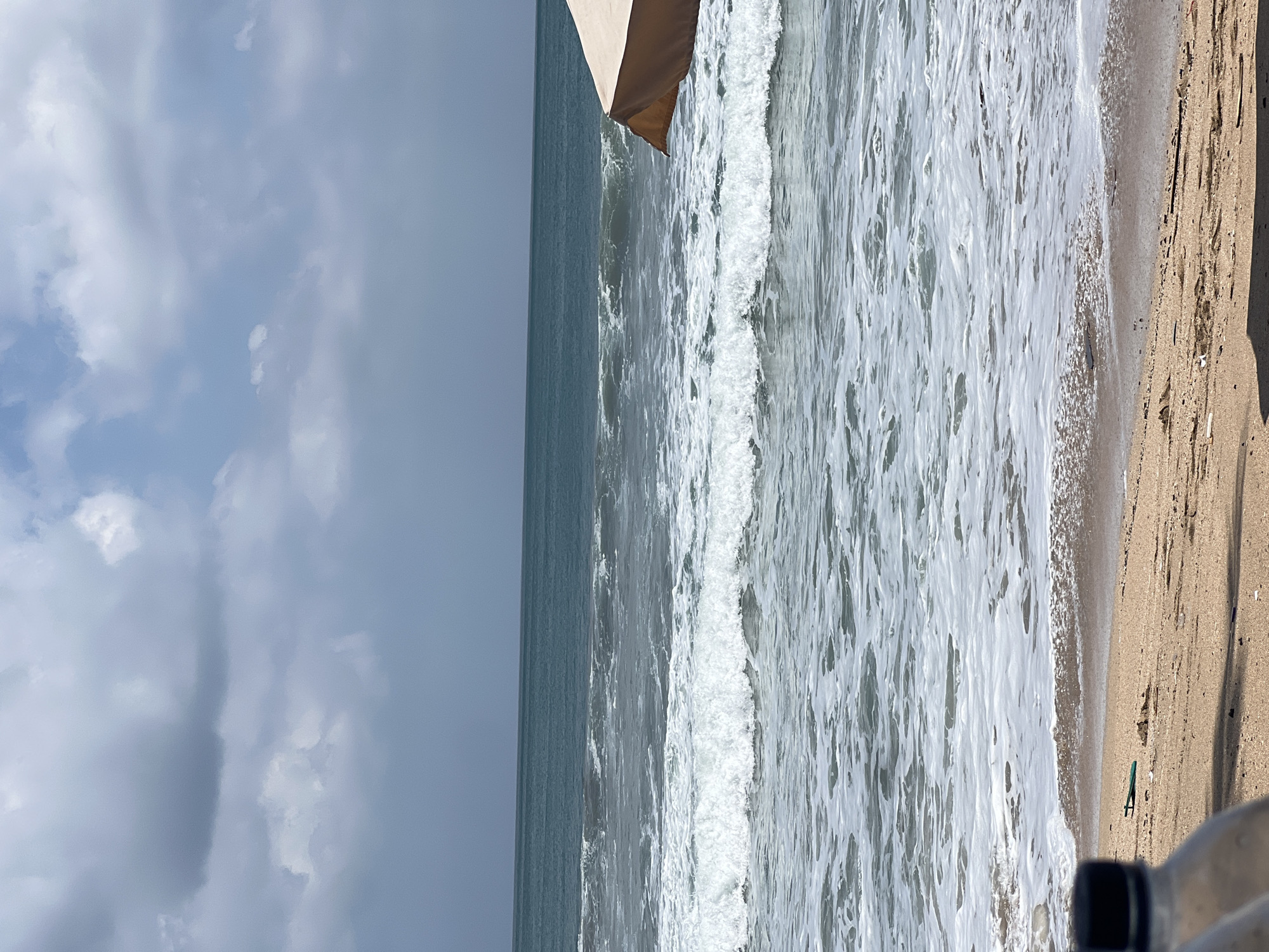 The image depicts a beach scene in Kuta, Indonesia. The ocean is visible with moderate waves and white foam reaching the shore. The sky is partly cloudy. In the foreground, a sandy beach is visible with some debris. Part of a tan beach umbrella is in the upper right corner of the frame. Two plastic bottles are partially visible in the lower left and bottom center of the frame. The overall environment suggests a typical beach setting.