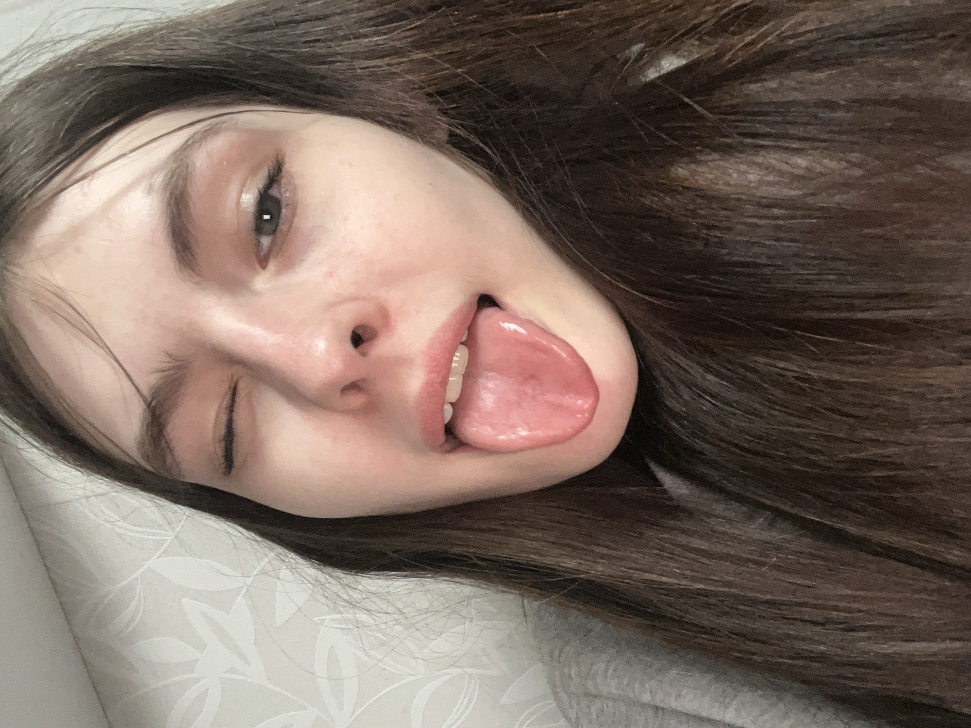 A young woman with long brown hair is pictured in a close-up selfie. She is lying down, possibly on a bed with a white patterned sheet. Her face is turned slightly towards the camera, and she is sticking her tongue out while winking one eye. The scene does not appear to be in Saint Petersburg, Russia as there are no indications of a geographical location.