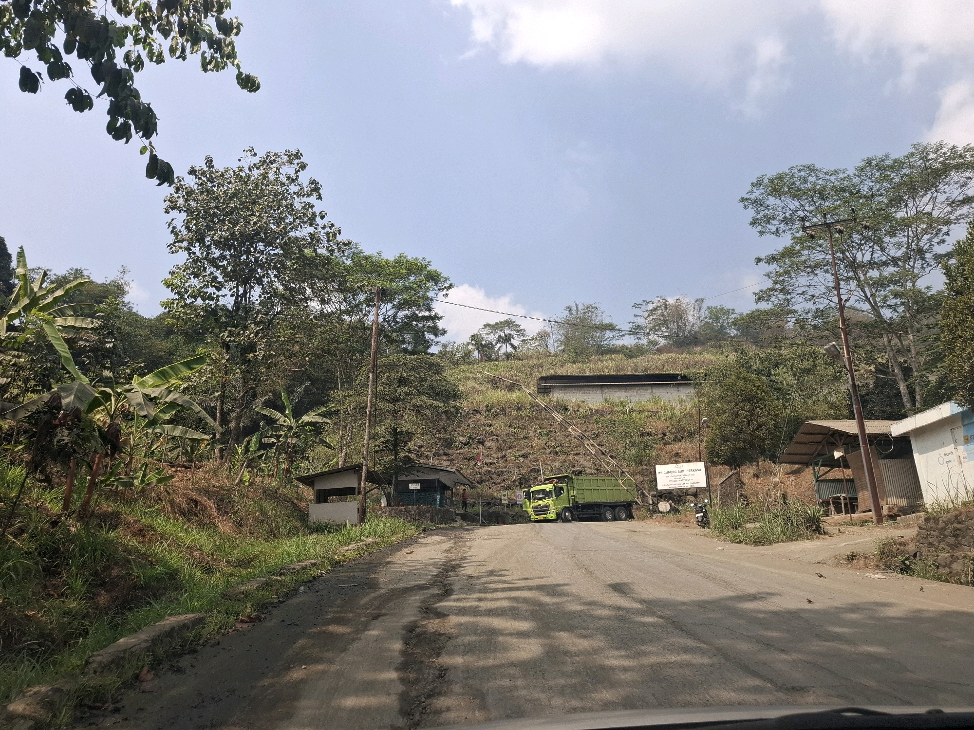The image shows a dirt road leading uphill in a tropical, mountainous region. On the left side of the road, there is lush green vegetation, including banana trees. Small, weathered shelters are situated along the road. Further up the road, a bright green dump truck is parked, facing towards the viewer. To the right of the truck, there's a building with a corrugated metal roof and a sign that reads "PT. GUNUNG BUMI PERKASA". The background consists of a steep, rocky hillside covered in trees and vegetation, with a long, low building visible at the top. The sky is clear with a few scattered clouds. The scene appears to be a mining or industrial site within a natural environment.