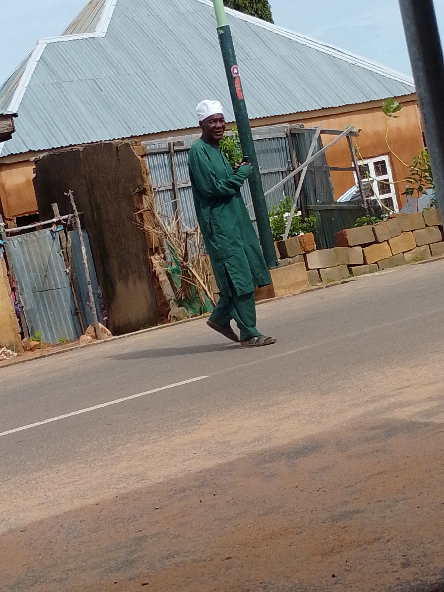 In Gombe, Nigeria, an elderly man wearing a green traditional outfit and a white cap walks along the side of a paved road. The man is holding a dark mobile phone in his left hand. Behind him, there is a building with a corrugated metal roof and brown walls. A pile of bricks is visible near the building, and to the left, a corrugated metal fence stands. The road has a white line marking its center. The environment appears to be a typical street scene in a Nigerian city.