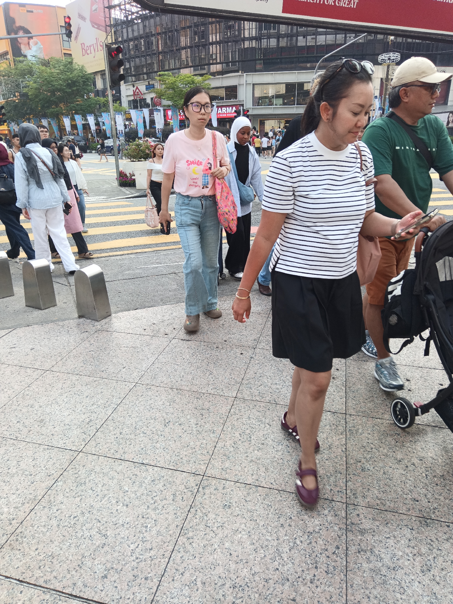 A medium shot captures a busy street scene in Kuala Lumpur, Malaysia, focusing on pedestrians crossing a crosswalk. In the foreground, a woman in a black and white striped shirt and black skirt walks away from the camera, her right hand holding a smartphone. To her right, a man in a green t-shirt and beige cap walks beside a baby stroller, also holding a smartphone in his right hand. Behind them, a woman wearing glasses and a pink t-shirt with a graphic print walks across the crosswalk. Further back, other pedestrians are visible, including a woman in a light blue striped shirt and white pants, and others in the background navigating the urban environment. The background features tall buildings, traffic lights, and various commercial signage, indicating a commercial district. The crosswalk is marked with yellow stripes on a gray road. The sidewalk is paved with granite tiles.