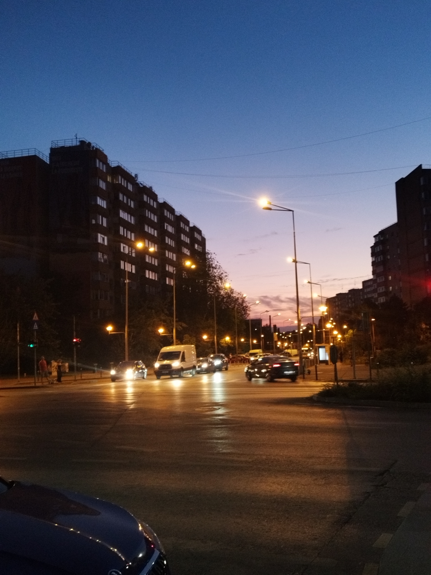 The image captures a street scene in Bucharest, Romania, at dusk. The sky is a deep blue transitioning to a soft purple and pink horizon, with scattered clouds. On the left, a large, dark residential building with many illuminated windows dominates the background, its facade featuring rows of balconies. Across the street, more buildings, also dark and with some lit windows, are visible, as are trees. The road in the foreground is wet, reflecting the bright headlights of passing vehicles and the warm glow of streetlights. Several cars are in motion, their headlights creating streaks of light due to the camera's exposure. A white van is clearly visible in the middle of the intersection, as is a dark sedan behind it. To the left of the road, a traffic light shows a green signal, and a few pedestrians are standing near it, seemingly waiting or conversing. The overall impression is of a busy urban environment transitioning into nighttime.