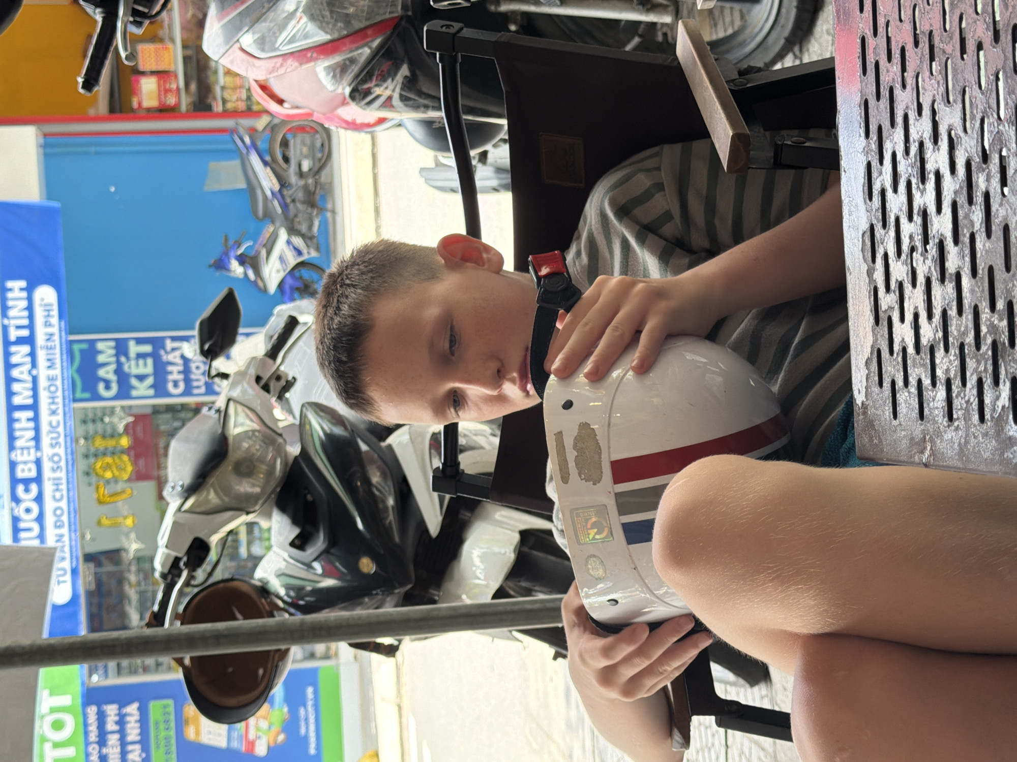 The image shows a young boy sitting in a chair, holding a white helmet with red and blue stripes. He is wearing a striped t-shirt. The background is blurred, but appears to be a street scene with motorcycles parked. There are signs with Vietnamese text in the background. This scene is likely in Đà Nẵng, Vietnam, given the presence of Vietnamese text and motorcycles commonly seen in Southeast Asian cities. The boy's expression is thoughtful or sad, as he gazes down at the helmet in his hands. The overall atmosphere is casual, suggesting a public space such as an outdoor cafe or waiting area.