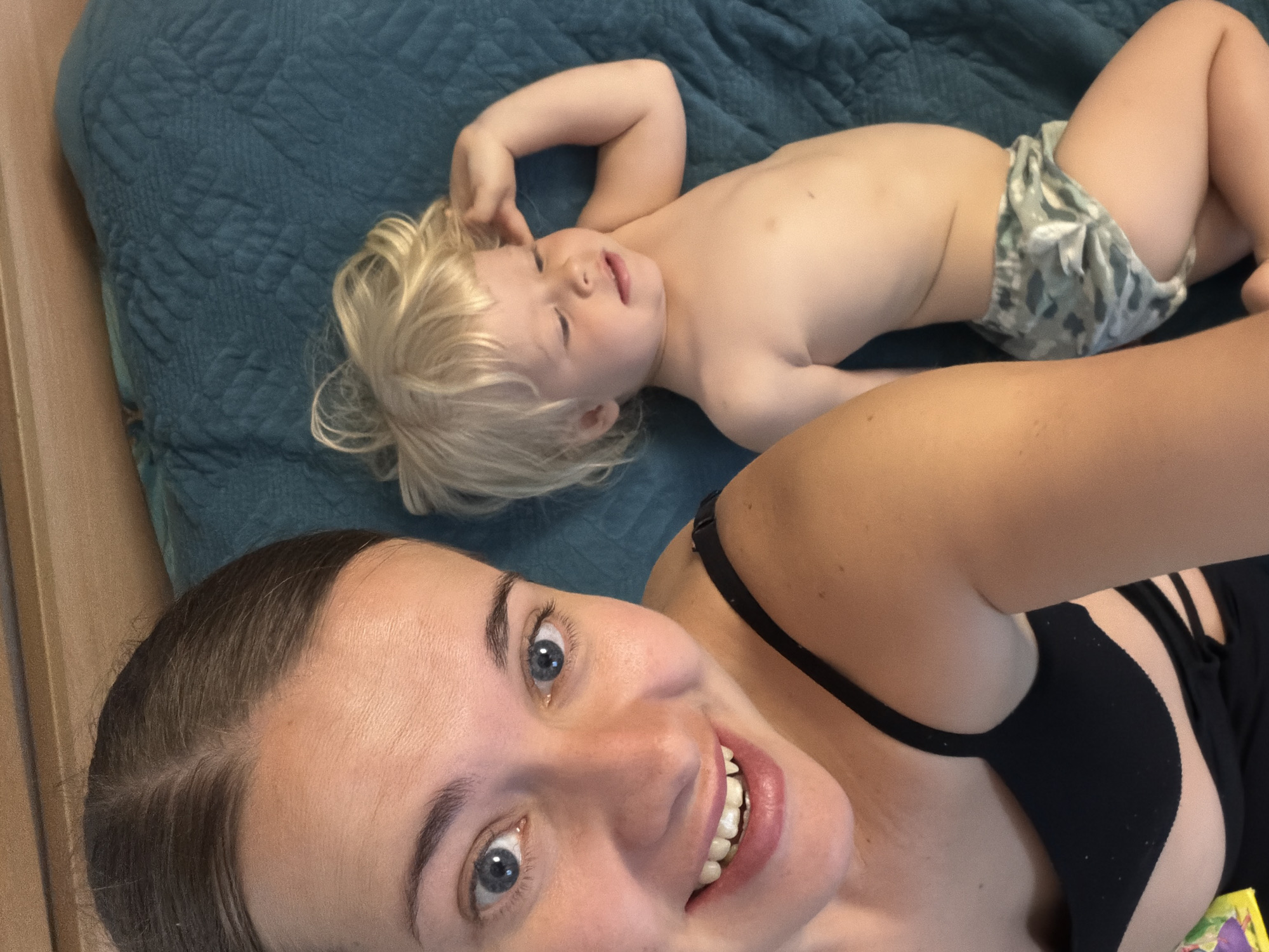 A selfie captures a woman with light skin, brown hair pulled back, blue eyes, and a smile exposing her teeth, lying down with a partially unclothed, blond-haired child on a teal-colored quilted surface. The child, wearing a diaper with a leaf pattern, is positioned behind and above the woman in the frame. The woman is wearing a black top. The location is unknown.