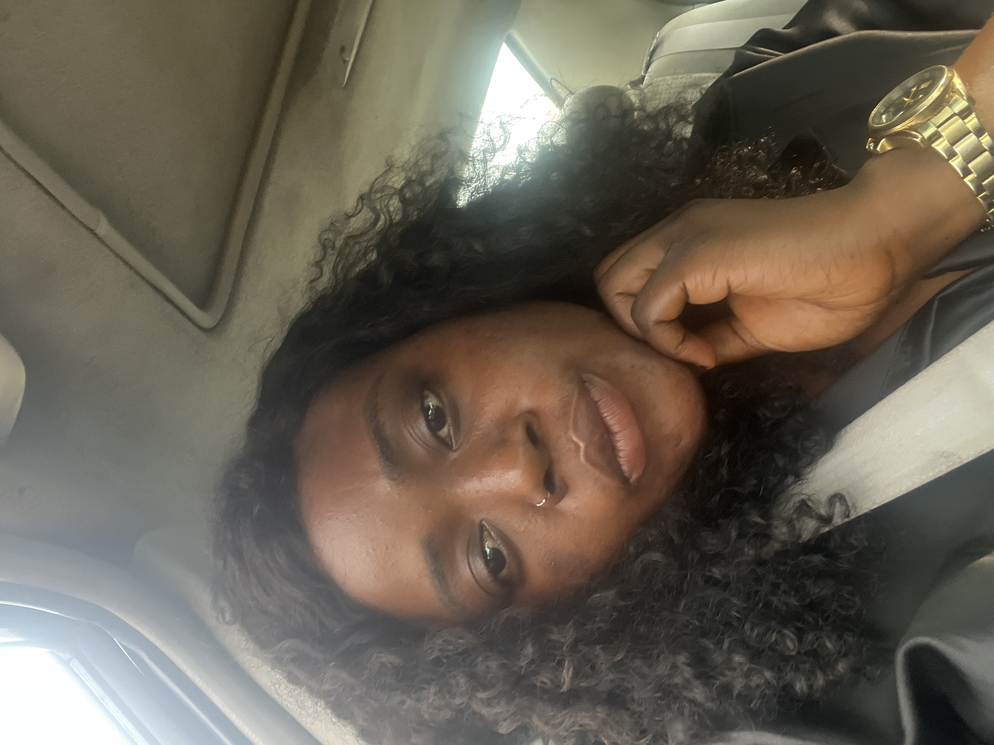 A close-up, slightly skewed selfie of a Black woman with dark curly hair is captured inside a vehicle, likely a car, identifiable by the interior roofing. She is wearing a gold wristwatch. The location, Abuja, Nigeria, cannot be determined from the image's visual information. No specific actions, interactions, or objects within the vehicle's interior are discernible other than the woman and the watch. The environment shows the car's ceiling and what appears to be a partially visible window, indicating daytime.