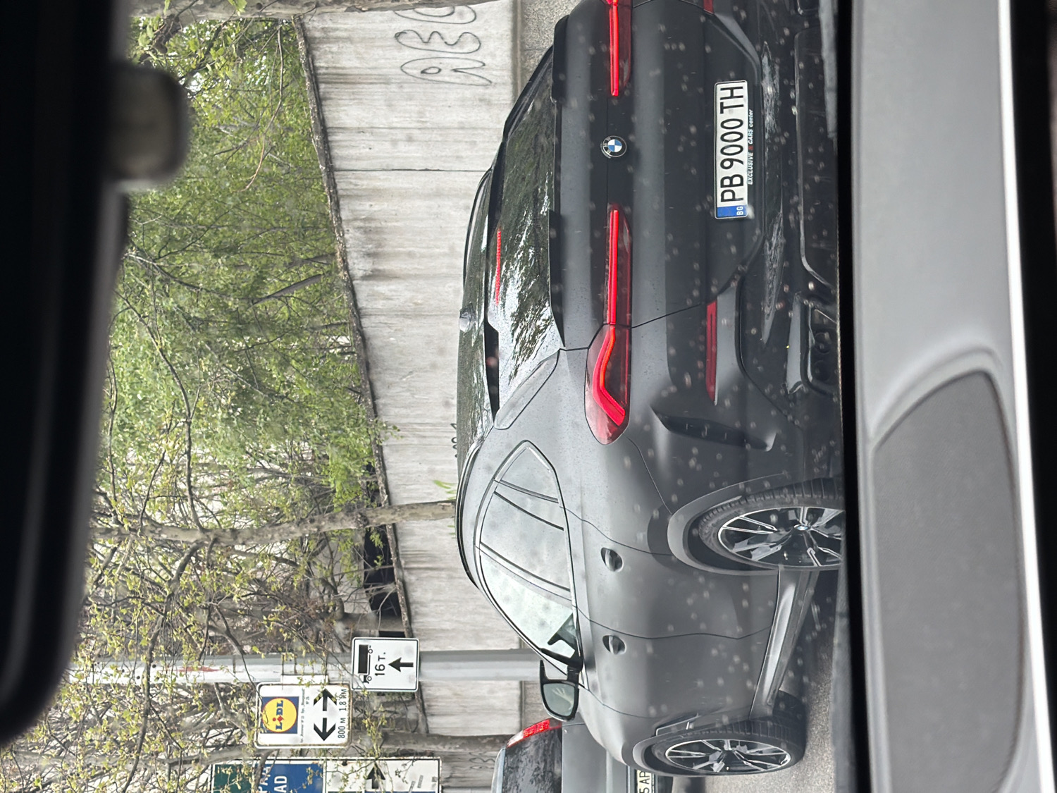 A dark grey BMW SUV, viewed from the rear, is the central subject of this image, captured through a window with numerous water droplets, suggesting recent rain or condensation. The vehicle bears a Bulgarian license plate "PB 9000 TH" with "BG" country code and "EXCLUSIVE CARS center" written below the number. The "PB" prefix indicates the Plovdiv region of Bulgaria.
To the left of the vehicle, green trees with light foliage are visible, along with several road signs. A prominent sign features the "LIDL" supermarket logo, with text "800 м 1.8 KM" indicating distances. Another sign shows "16 т." (16 tons) with an upward arrow, likely denoting a weight limit. Parts of other signs with text like "AD" and "5 AP" are also discernible. In the background, a concrete wall has partially visible graffiti that reads "ABC". The overall scene appears to be an urban or suburban setting, possibly alongside a road or underpass. The image is taken during the daytime.