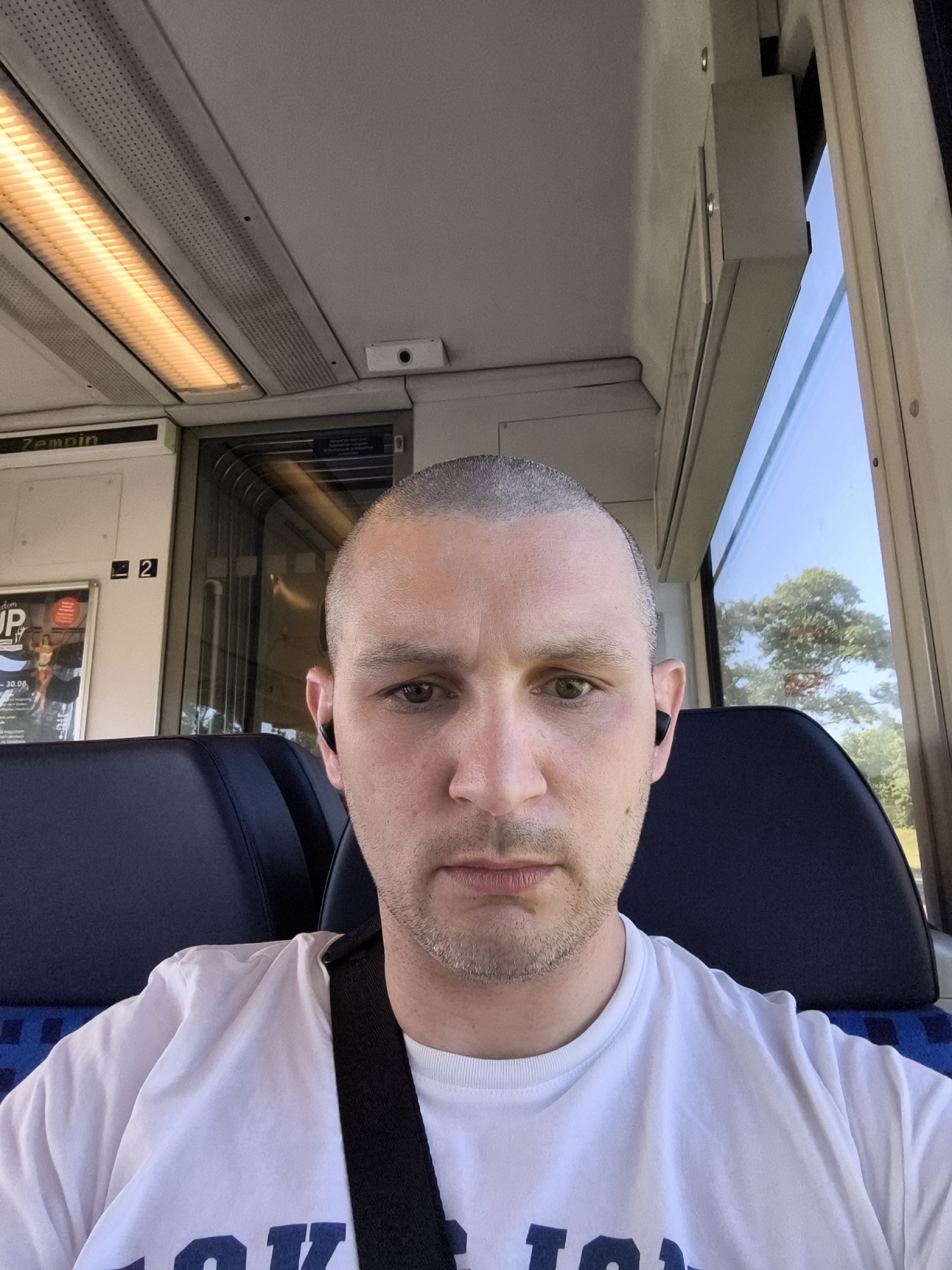 The media file is a selfie taken by a person with a shaved head, wearing a white t-shirt and black earbuds, while sitting inside a train carriage. The person is looking directly at the camera with a neutral expression.
The setting is clearly a train with blue seats visible. Above the person's head, fluorescent lights are mounted on the ceiling, indicating the interior of the train. A window on the right side of the frame shows trees and a blue sky, suggesting it's daytime and possibly sunny weather outside. The interior of the train appears to be in good condition, with a poster visible on the wall behind the person's left shoulder.
Visible text includes "Zemoin" written on a sign above the poster. The poster itself has some text that is partially obscured, but "UP" and "30.08." are discernible, possibly indicating an event or advertisement with a date. There is also a number "2" visible on the wall.
The overall mood is ordinary, depicting a person on a train journey during the day. The location context of Koserow, Germany, suggests the train is likely operating in that region. There are no other people visible in the frame. The focus is entirely on the individual and their immediate surroundings within the train.