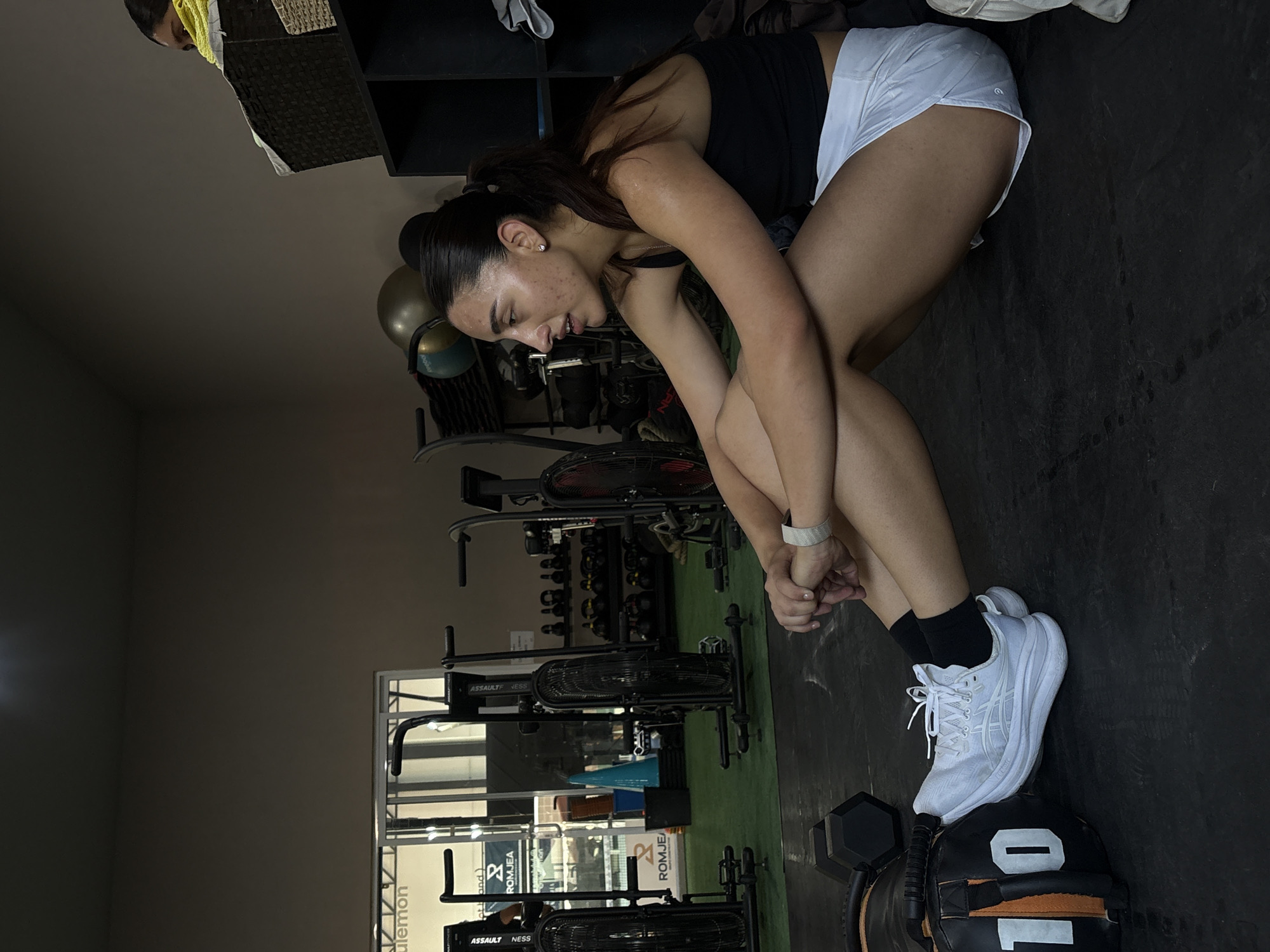 A young woman is seated in a gym in San Bernardino Tlaxcalancingo, Mexico. She is wearing a black tank top and white shorts. She is resting her feet on a black and orange weight bag, and wearing white sneakers and black socks. She is resting with her arms on her knees. Gym equipment, including stationary bikes, weights, and a rack, are visible in the background. A window is visible in the background too.