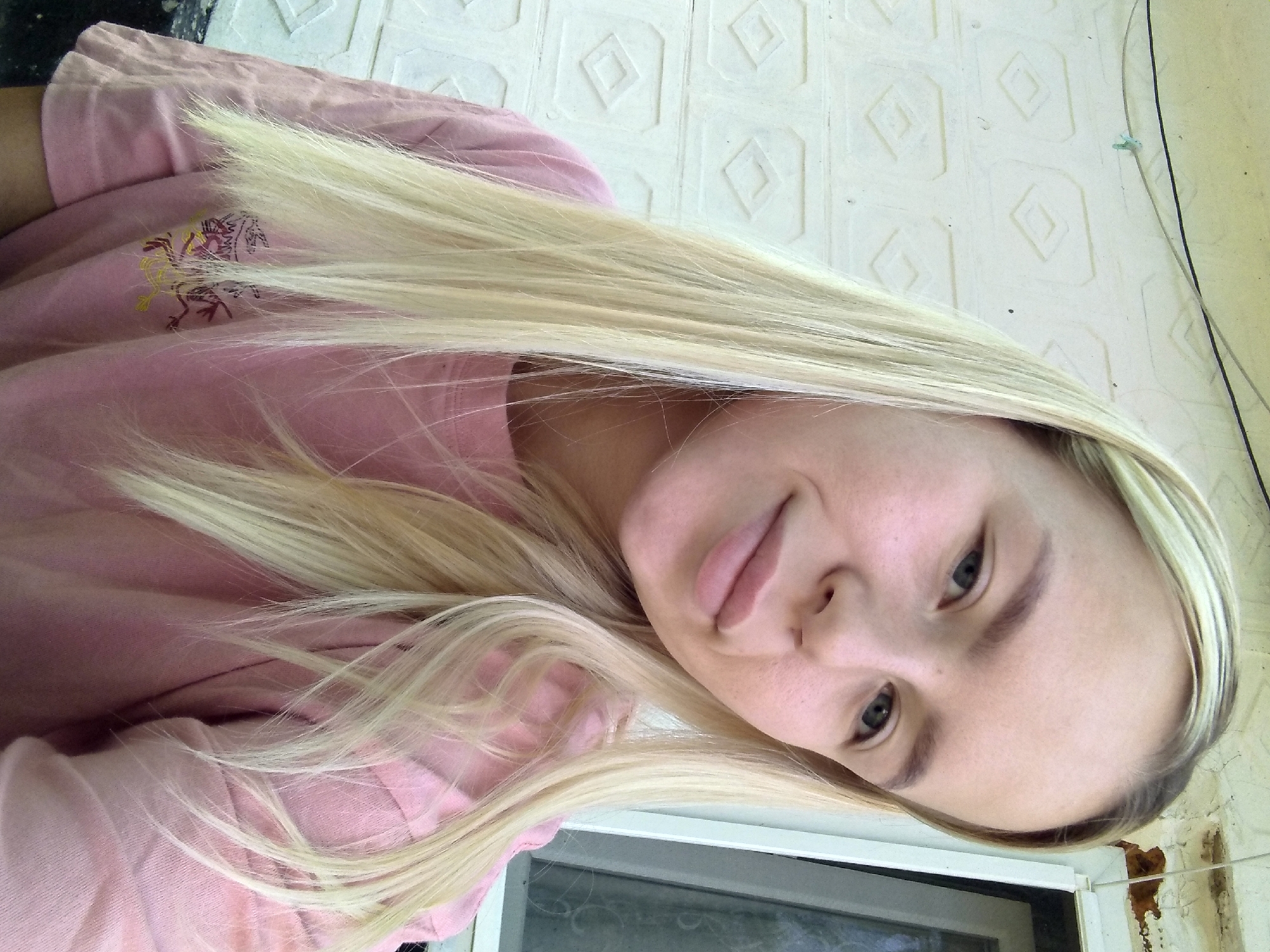 A young woman with long blonde hair is pictured from the chest up, in a selfie. She is wearing a pink t-shirt. The background consists of a white textured wall with a diamond pattern and a window. The image appears to be taken outdoors. The location is identified as Radisne, Ukraine.