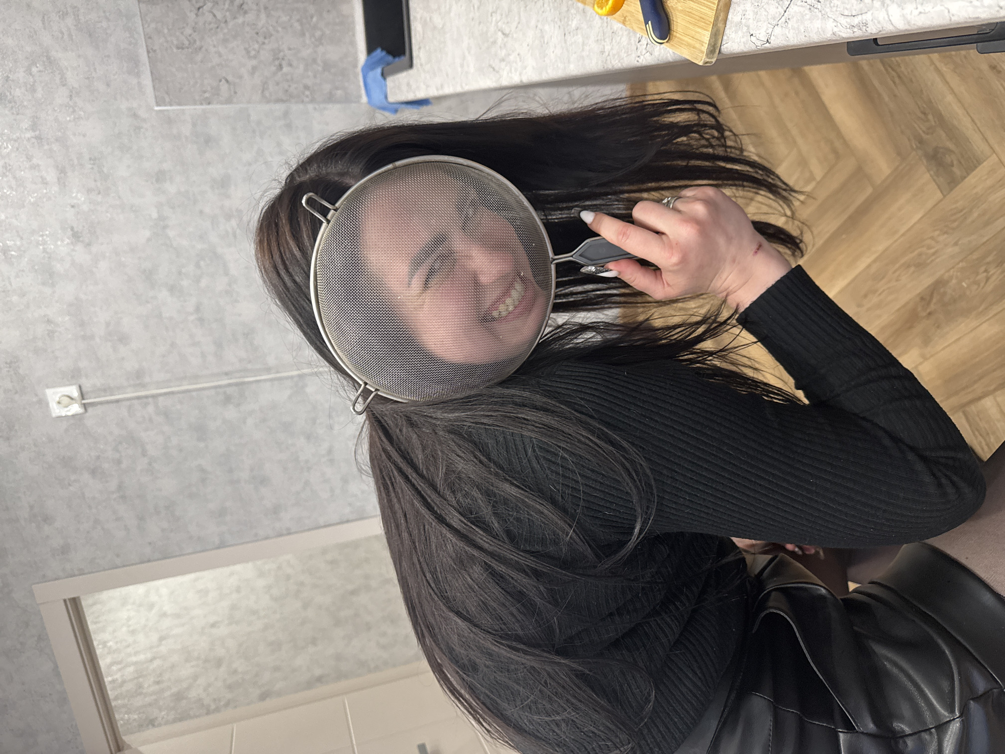The image captures a playful indoor scene, likely set in a kitchen. The primary subject is a woman with long, dark hair, wearing a black ribbed long-sleeved top and a black, possibly faux-leather, skirt. She is holding a round metal mesh kitchen strainer in front of her face, through which her smiling expression is clearly visible. Her wide, toothy smile conveys a sense of amusement and joy. Her left hand, adorned with a ring and white nail polish, grips the handle of the strainer. A small red mark or scratch is noticeable on her left wrist.
Her long, dark hair cascades around her head and spreads across surfaces. In the background, a light gray countertop is visible, hosting a wooden cutting board with a sliced orange and a knife. A blue item, possibly a cleaning cloth or sponge, is also present on the counter. The wall behind her features a textured light gray surface with a white power outlet and a corresponding cable. Further back, a frosted window or glass-paneled door is discernible. The floor, visible in the lower part of the image, is light brown with a wood-like pattern. The lighting suggests an indoor setting, well-illuminated. There is no visible text in the image. The entire image is rotated 90 degrees clockwise from what would be a natural upright orientation.