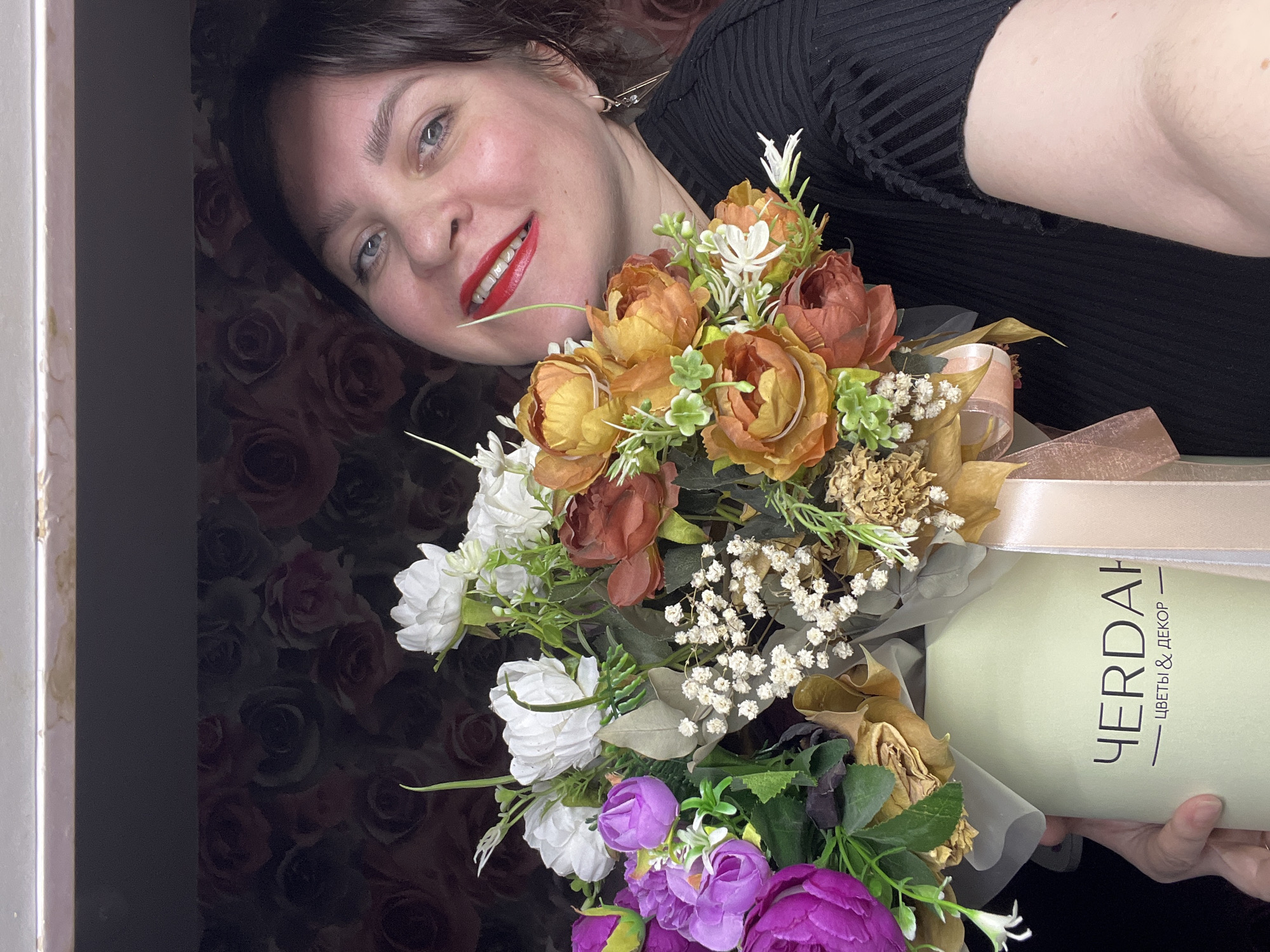 An adult female is depicted in a close-up indoor shot, holding a large artificial flower bouquet in a cylindrical box. The setting is identified as the city of Noviy, Russia, within an interior space featuring dark wallpaper patterned with red and black roses. A narrow, light-colored horizontal surface is visible along the top edge of the frame. The woman, positioned to the right of the bouquet, smiles broadly at the camera, displaying red lipstick, blue eyes, and dark hair. She wears a black ribbed top, and a geometric earring is visible on her left ear. She holds the light green cylindrical box, which bears the Cyrillic inscription "ЧЕРДАК" (CHERDAK) above "ЦВЕТЫ & ДЕКОР" (FLOWERS & DECOR), and is adorned with a light beige ribbon. The bouquet itself comprises various artificial flowers in dominant shades of white, purple, orange, rust-brown, and pale yellow, interspersed with green foliage and small white accent flowers. Several petals exhibit signs of browning or discoloration.
