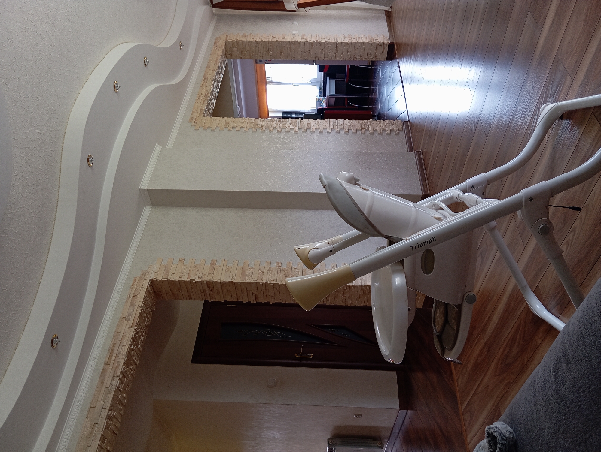 In the city of Hlynytsi, Ukraine, an indoor scene captures a modern living space. The room features a curved ceiling with recessed lighting, complemented by decorative plasterwork and stone veneer accents around archways. The flooring is polished hardwood. In the foreground, a white high chair with a beige seat is positioned, facing towards the viewer. A dark wooden door is visible in the left background, and a doorway to another room is in the right background, revealing a glimpse of a dining area with a table and chairs. The scene appears to be a domestic environment, possibly a dining or living area.