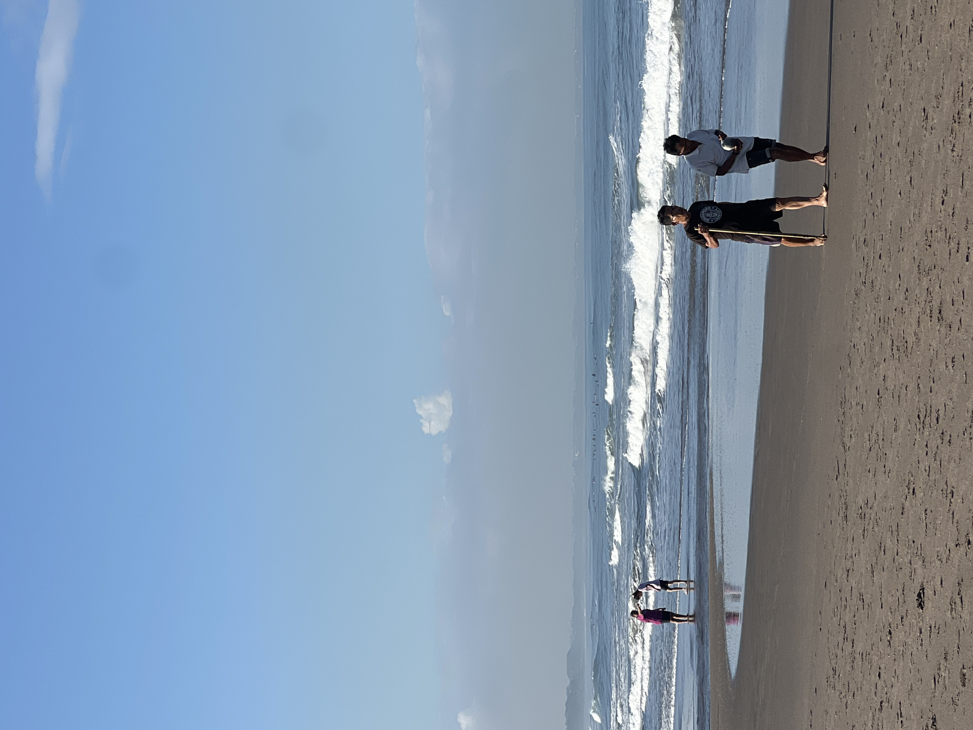 In North Kuta, Indonesia, two men walk on a sandy beach under a clear blue sky with a few wispy clouds. The man on the left wears a black t-shirt and dark shorts, holding a wooden stick. The man on the right wears a white polo shirt and dark shorts, carrying a round object. In the shallow water behind them, two individuals are visible, one wearing a pink garment and the other white. The ocean has moderate waves crashing onto the shore. Distant landmass and surfers are faintly visible on the horizon.