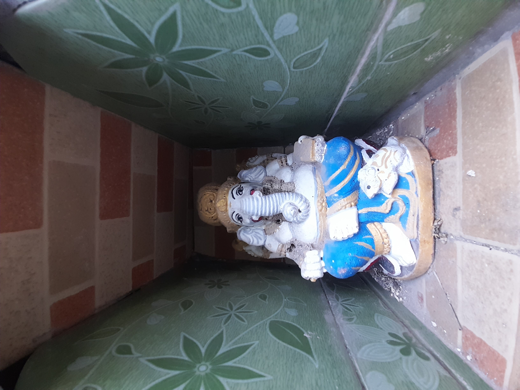 The image presents a top-down, close-up view of a statue of Lord Ganesha, positioned within a narrow corner or alcove. The statue is primarily white, adorned with striking blue and gold clothing or decorative elements, and features a golden crown on its head. Its elephant trunk is curled to the left, and subtle red detailing is visible on its lips. There is a noticeable amount of dirt and small debris accumulated around the base and on parts of the statue.
The immediate surroundings consist of two different patterned surfaces. The surface on the left, which appears to be a wall or floor depending on orientation, is covered in a green material, possibly linoleum or wallpaper, displaying a repeating pattern of lighter green floral and leaf motifs against a darker green background. The vertical wall on the right is covered with rectangular tiles in alternating shades of light brown and reddish-brown. The statue rests on a light-colored, possibly beige-tiled surface.
The scene depicts a static arrangement, likely a devotional display. The lighting suggests it is daytime, with soft, diffused light illuminating the area. The combination of decorative wall coverings implies an indoor setting, possibly a household shrine or a dedicated niche within a building. No visible text is present in the image.