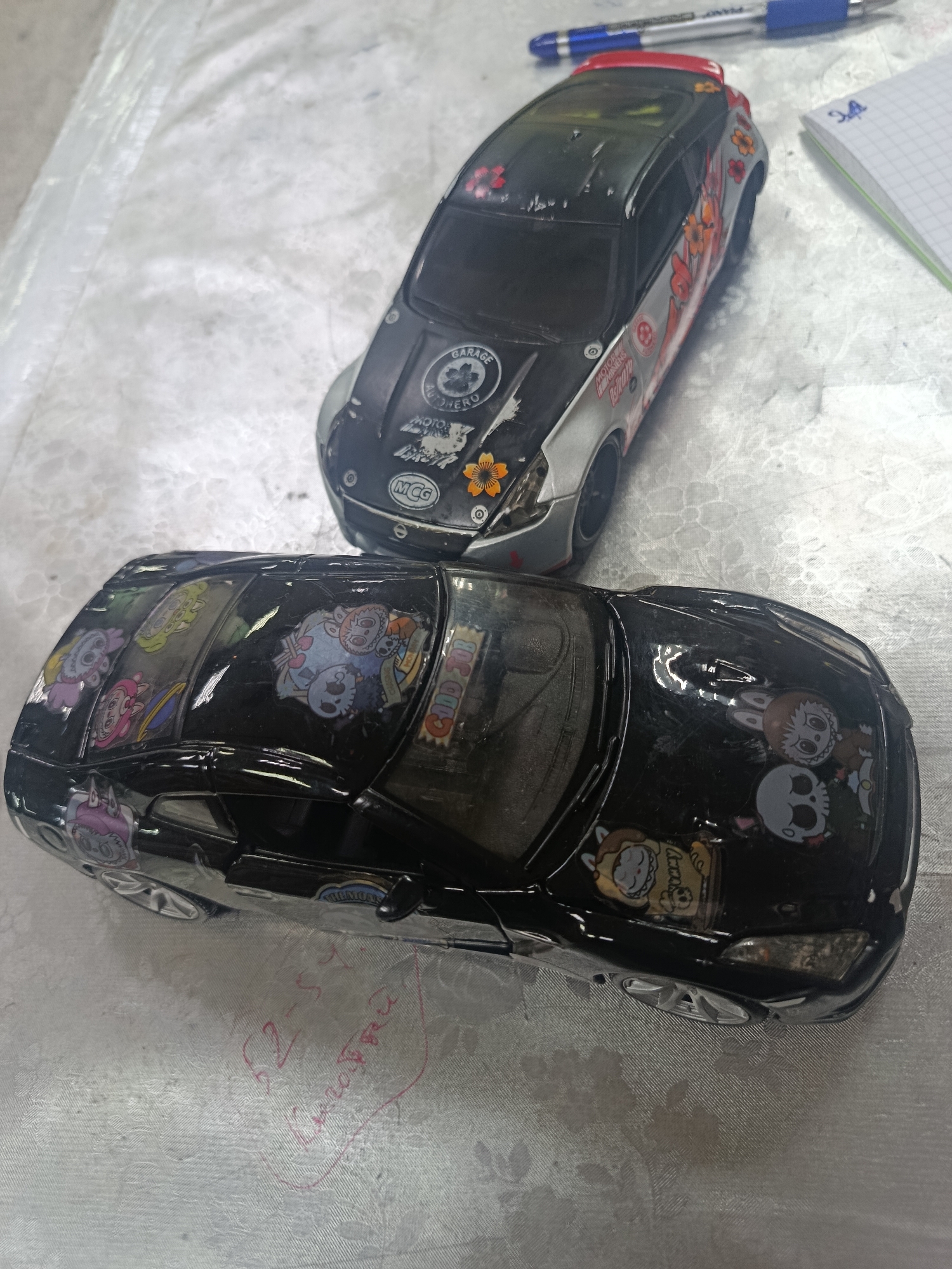 Two toy cars are positioned on a white tablecloth. The car in the foreground is black and adorned with cartoon stickers. The car in the background is gray and silver, also decorated with various stickers and decals. A blue and red pen lies in the upper right portion of the image, next to a small, open notebook with a grid pattern. The environment appears to be indoors.