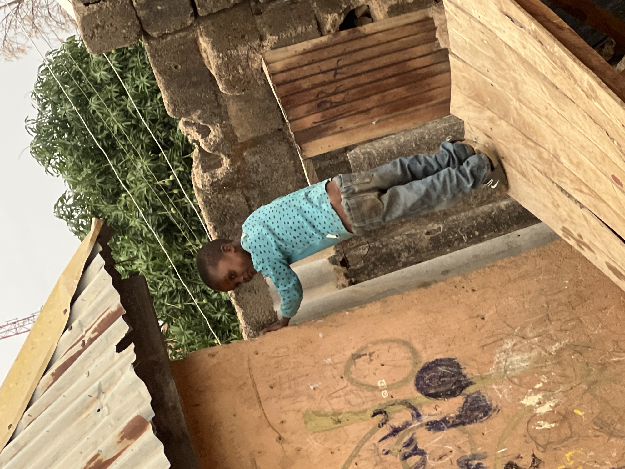 A child in a long-sleeved blue shirt with small white polka dots and denim pants performs a push-up on a weathered, painted surface, likely a rooftop or elevated platform. His feet are planted against a stack of wooden planks. A building constructed of tan blocks is adjacent to him, with vegetation and a metal roof visible in the background. Based on common architectural styles and environmental features, the location is likely in an urban area within the city of Jos, Nigeria.