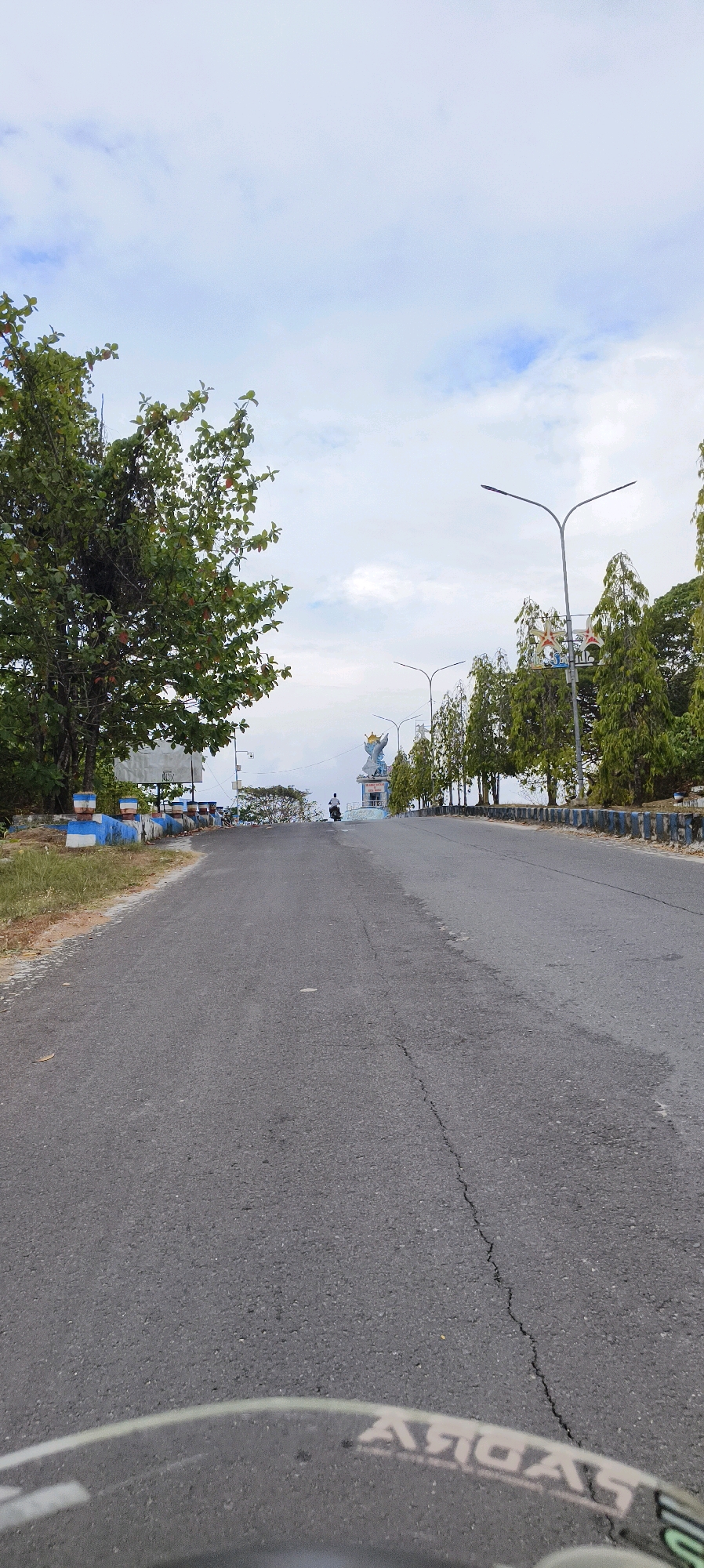The image shows a road leading uphill towards a statue of a mermaid with a crown. The road is paved and cracked in places. On the left side of the road, there is a tree with green leaves and a short wall with blue and white stripes. On the right side of the road, there are streetlights and more trees. In the distance, there is a person riding a motorcycle. The sky is cloudy. This location is identified as the entrance to Waisai, Raja Ampat, Indonesia.