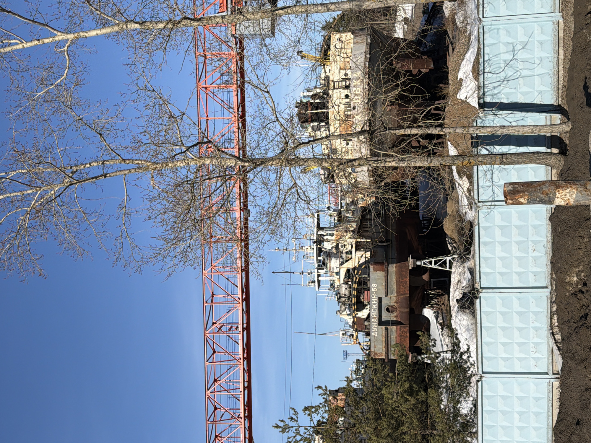 This image depicts an industrial scene in Chaikovsky, Russia, under a clear blue sky. In the mid-ground, a large, multi-deck industrial vessel, identified by the Cyrillic text "ДУНАЙСКИЙ-58" (Dunayskiy-58) on its stern, is positioned out of water, supported by white metal structures. The vessel exhibits extensive rust and weathered white and grey paint. Behind it, parts of at least one other similar vessel are visible. Overarching the scene in the upper portion of the image is a prominent red metal truss structure, likely part of a gantry crane or an overhead walkway. In the foreground, a light blue concrete fence with a raised diamond pattern spans the width of the frame. Behind the fence, patches of melting snow are visible on uneven ground. The immediate foreground consists of dark, disturbed earth. Several bare deciduous trees with thin branches are scattered throughout the mid-ground, partially obscuring the vessels. Evergreen trees are present on the far left. No individuals or active movements are discernible within the frame.
