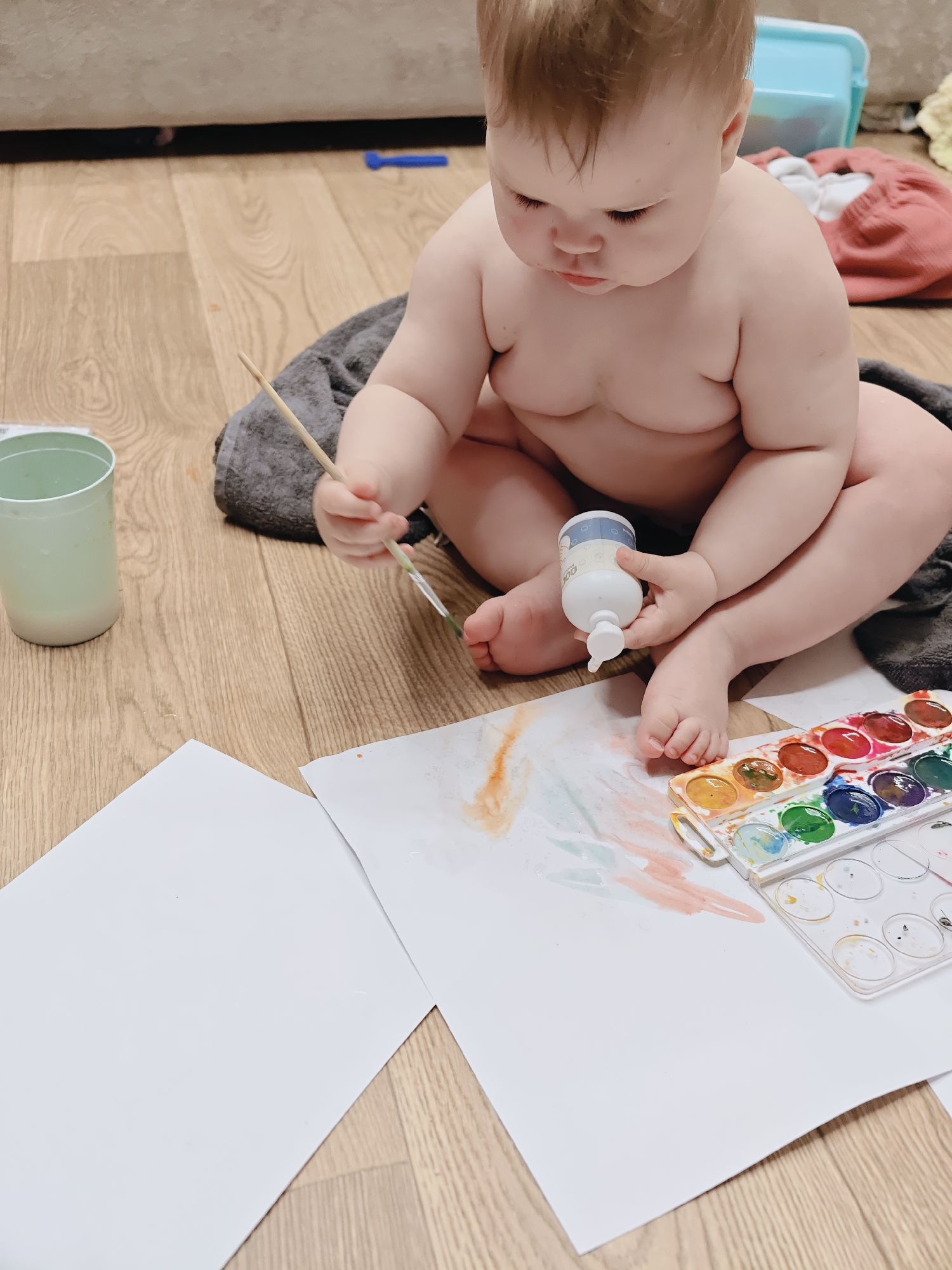 A baby is sitting cross-legged on a gray towel placed on a wooden floor. The baby is holding a paintbrush in their right hand and a white bottle in their left hand. In front of the baby are two sheets of white paper, one blank and the other with some paint on it, and a set of watercolor paints. A light green cup is visible to the left of the baby. In the background, a light blue storage container and some clothing can be seen. The scene is likely indoors in the city of Chaikovsky, Russia.