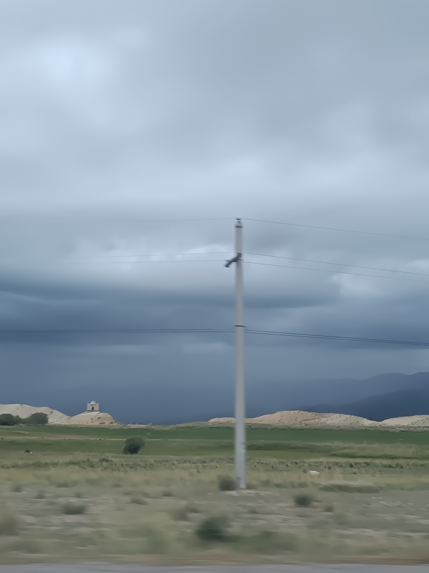 The image shows a landscape with a cloudy sky. In the foreground, there is a light-colored utility pole with power lines running across it. The middle ground features rolling hills with patches of green grass and dry brush. On one of the hills, a small white structure with a dome is visible. In the distance, dark mountains are silhouetted against the sky. The scene appears to be in a rural or semi-rural area. The location is identified as Sasyk-Bulak, Kyrgyzstan.