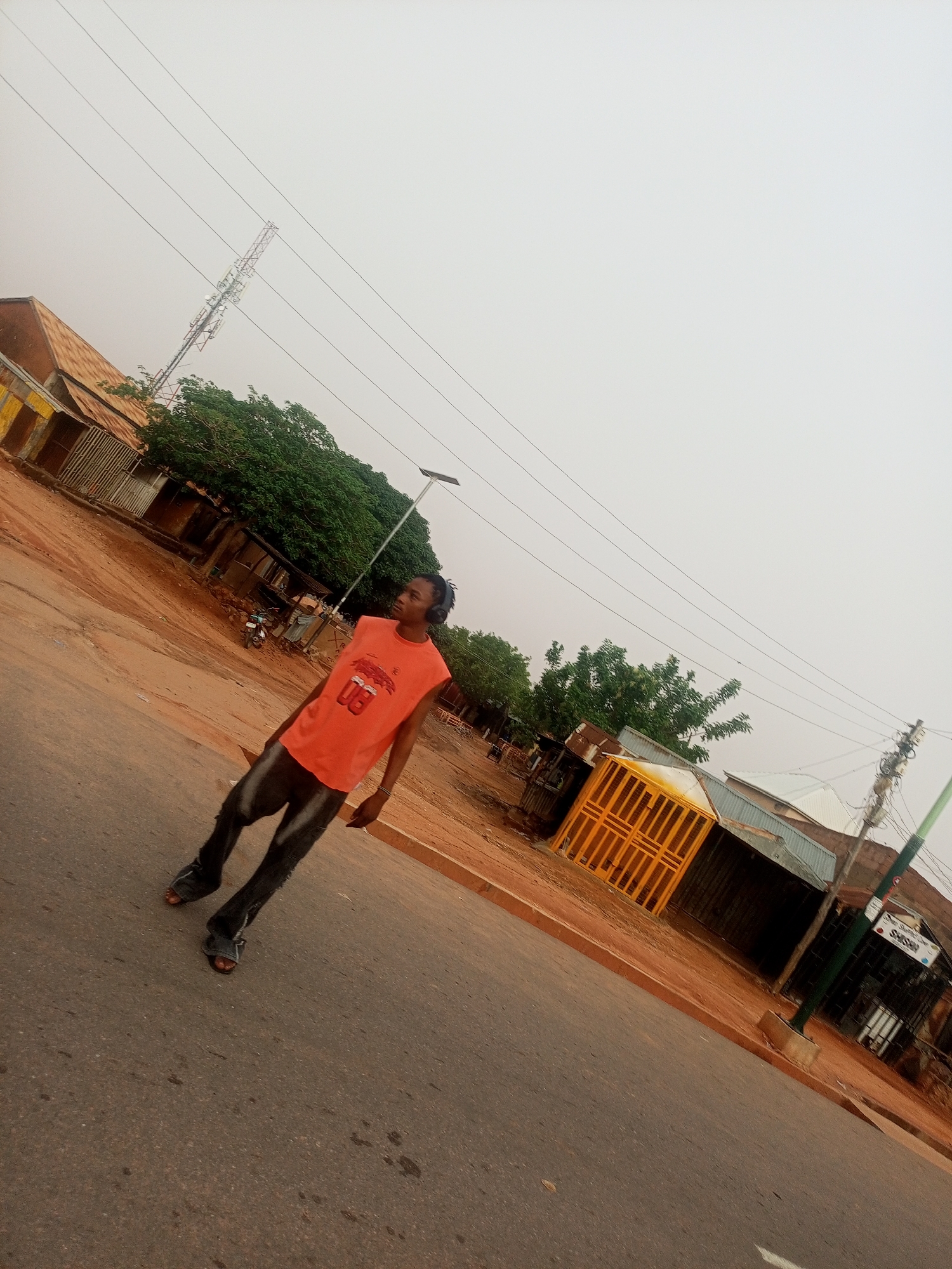 A young man is standing on a paved street in Tempure, Nigeria. He is wearing a sleeveless orange shirt, dark trousers, and sandals. He has short hair and is wearing headphones. The street appears to be in a residential or commercial area, with several buildings visible in the background. There is a cell tower in the background. The sky is overcast.