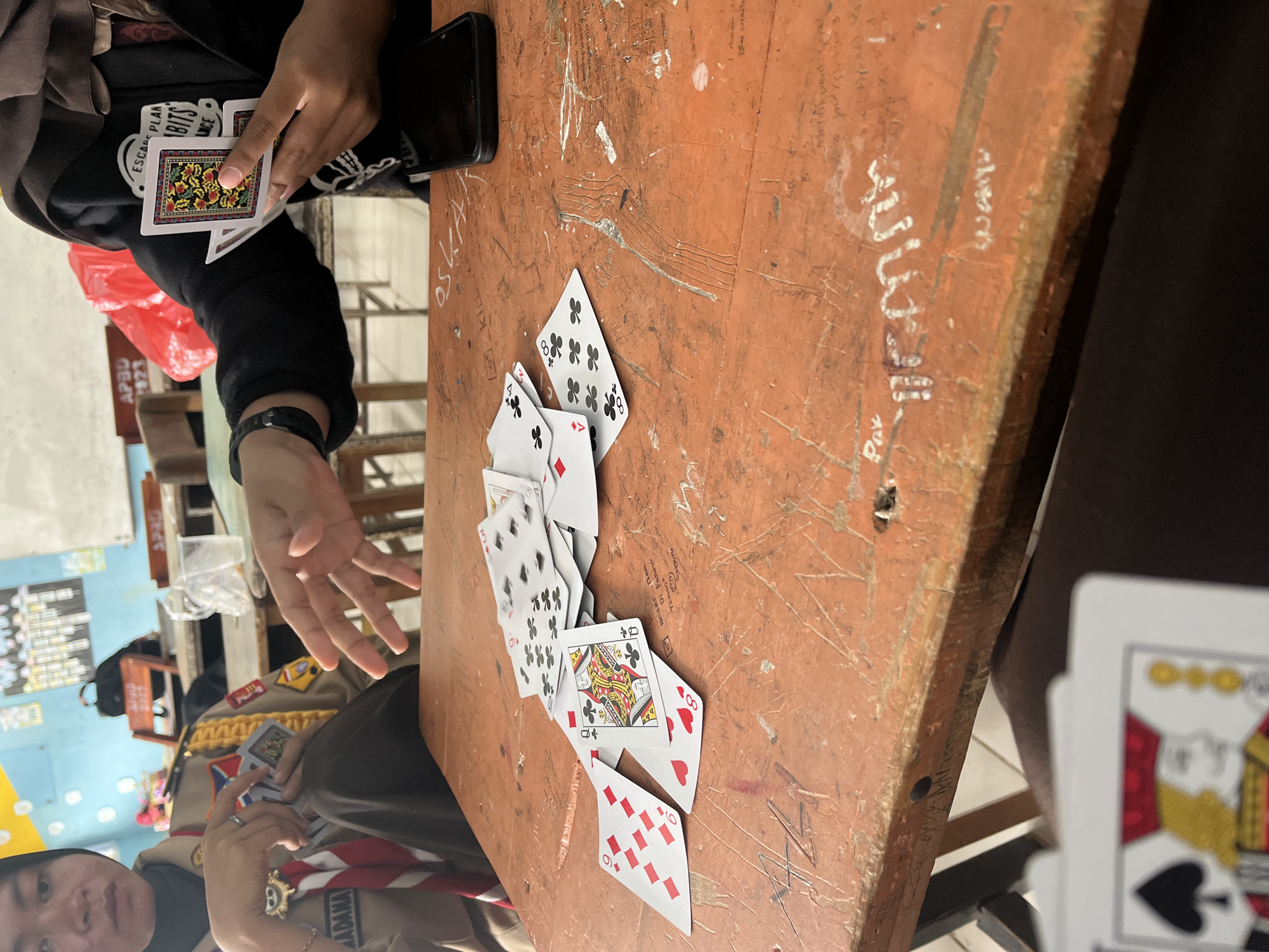 The image shows a close-up view of an orange wooden table where several playing cards are scattered. Two people are visible, partially cropped. One person, on the left, is wearing a scout uniform with a red and white scarf. Their hands are holding a stack of cards. The other person, on the right, is wearing a black jacket with a hijab and is also holding cards, with their hand extended over the table. The table has various markings and graffiti. In the background, there are classroom elements such as a whiteboard and chairs, suggesting the location is likely a school. The image appears to be taken in Pompanua, Indonesia, as indicated by the context.
