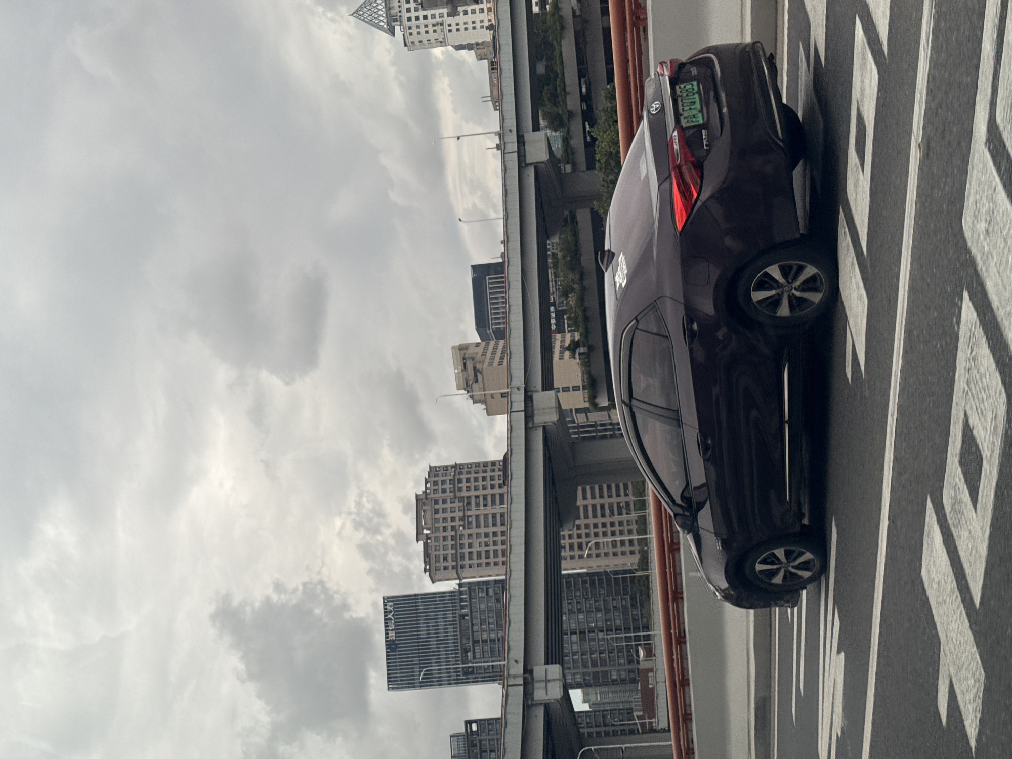A dark red sedan drives on an elevated highway in Shanghai, China. The car is in the right lane of a multi-lane highway, with white markings on the asphalt indicating traffic lanes and a crosswalk. The sky is overcast, with scattered clouds. In the background, a cityscape of tall buildings and overpasses is visible. The car's license plate reads "FA FQ7685".
