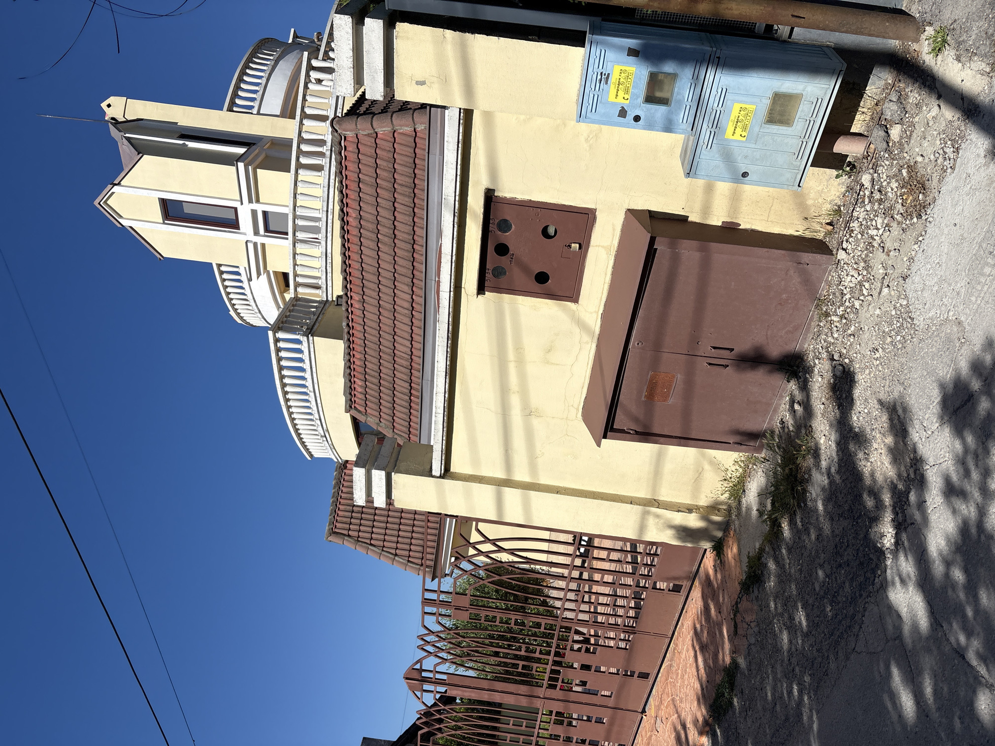 The image depicts a section of a residential building in Varna, Bulgaria, on a sunny day. The building features a prominent yellow facade, a terracotta tiled roof, and a distinctive curved white balustrade on an upper level. To the left of the building is a large, ornate brown metal gate with an intricate geometric design, partially obscuring some greenery behind it. In front of the building's wall, there are two brown metal boxes and two light blue utility cabinets stacked vertically. The ground in front of the building is a mixture of cracked asphalt, gravel, and sparse vegetation, with shadows from overhead foliage cast across the scene. The sky is a clear, bright blue. No people are visible in the image.