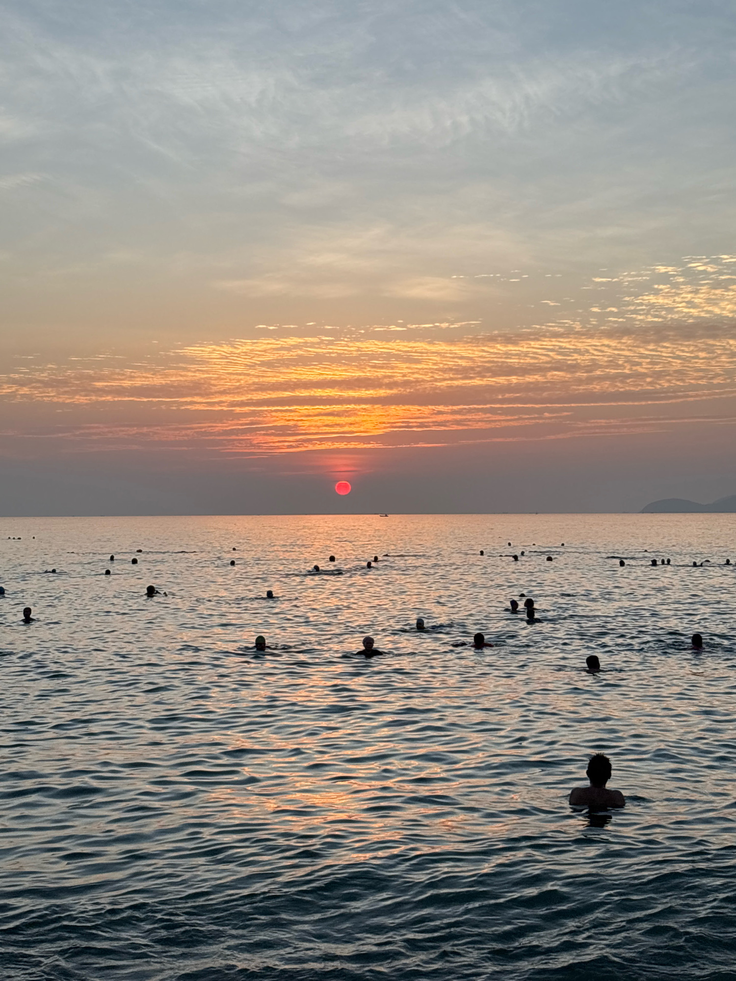 The image depicts a wide shot of Nha Trang Bay in Vietnam at sunset. The sky is filled with a gradient of soft blues and oranges, with wispy clouds catching the last rays of sunlight. A vibrant red sun is setting near the horizon, casting a warm glow across the scene. The sea is calm, with gentle ripples reflecting the colors of the sky. Numerous people are visible in the water, some partially submerged, appearing to be swimming or enjoying the evening. They are scattered across the bay, with no apparent interactions between them. The overall impression is one of a peaceful and serene sunset on a popular beach in Nha Trang.