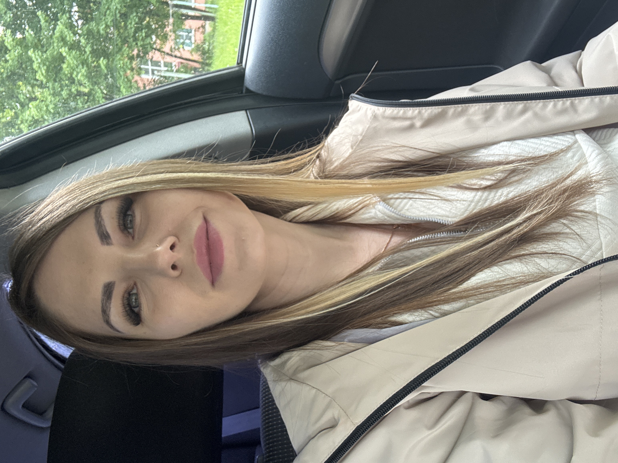 A woman with blonde and brown hair sits inside a car. She is wearing a light tan jacket and has pink lipstick. The car's interior is visible with a black headliner, door panel, and seat. Outside the car window, green trees and part of a building are visible. The photograph does not contain enough information to establish the exact location.